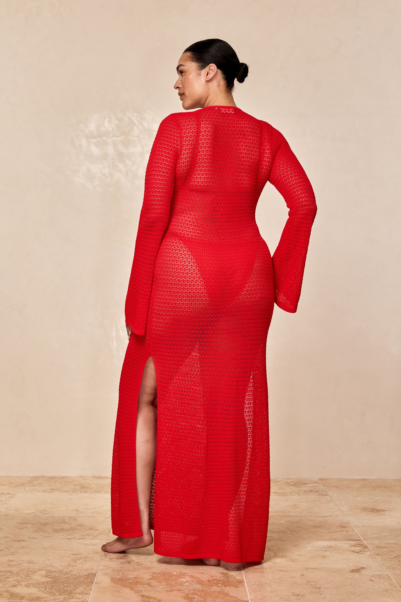 Rear view of a woman wearing a long red crochet dress with a thigh slit, barefoot, hair in a bun, neutral background.