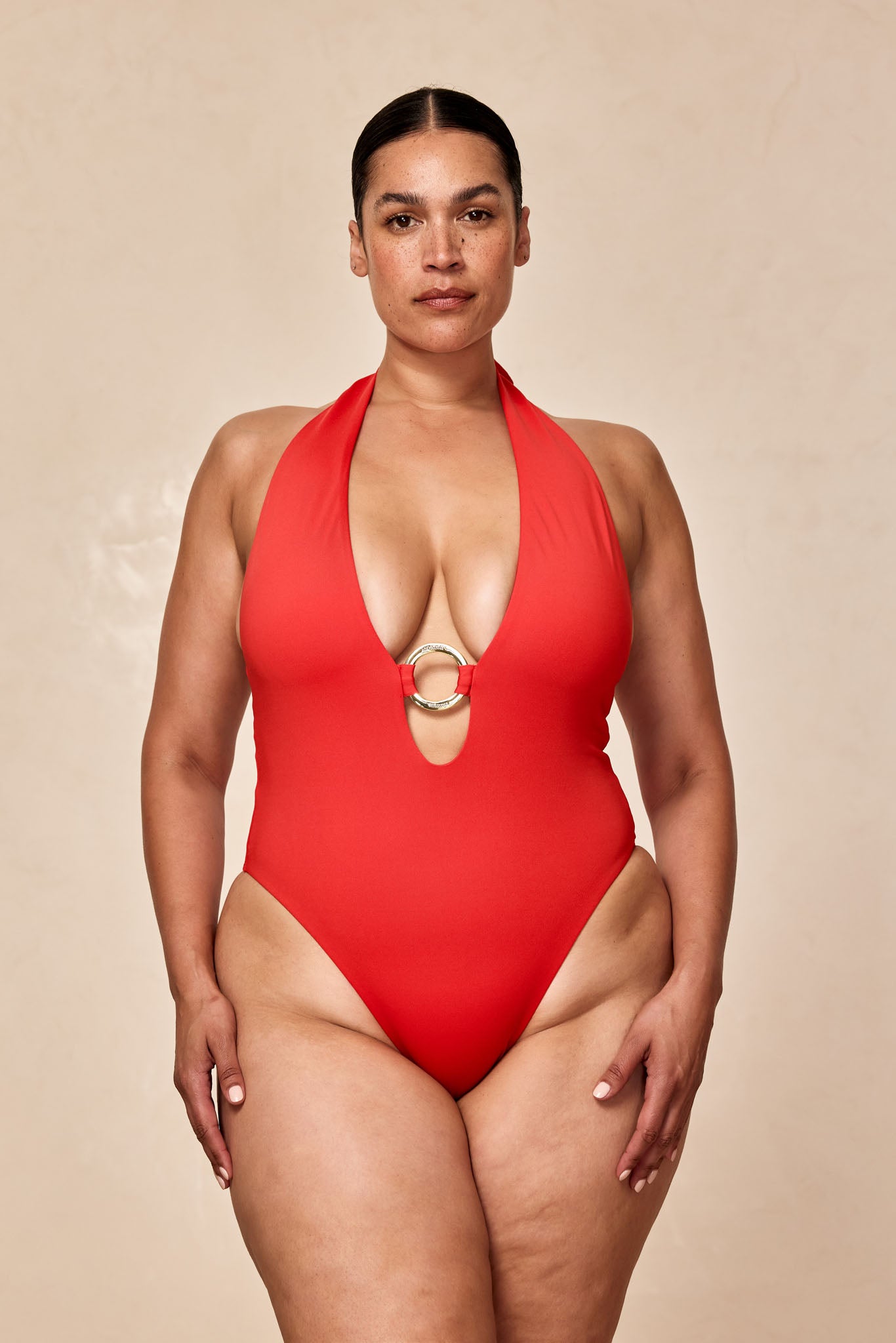 Woman wearing red halter one-piece swimsuit with deep V neckline and gold center ring, standing against a neutral background.