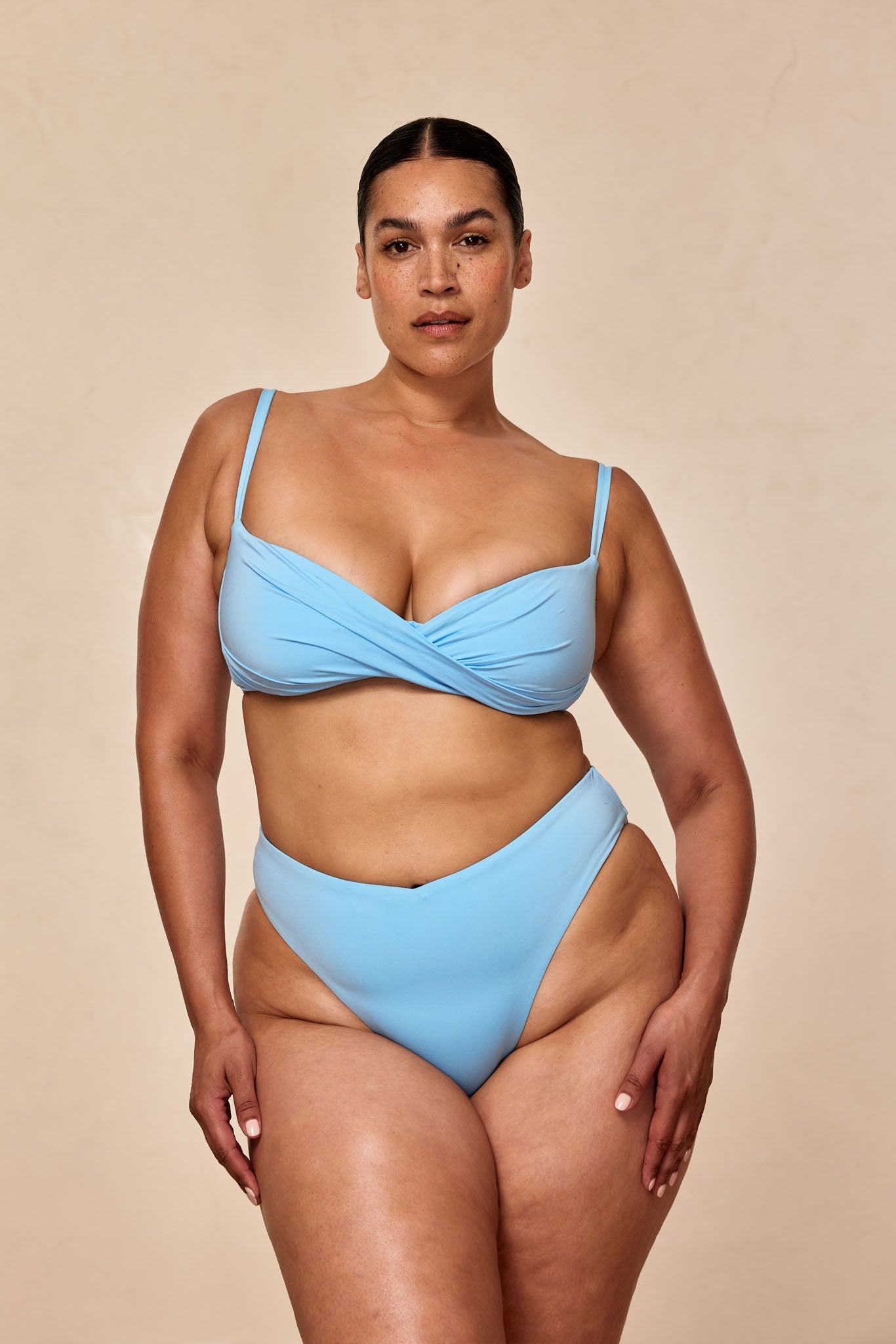 Curvy model wearing a light blue bikini set, front view against a neutral beige backdrop