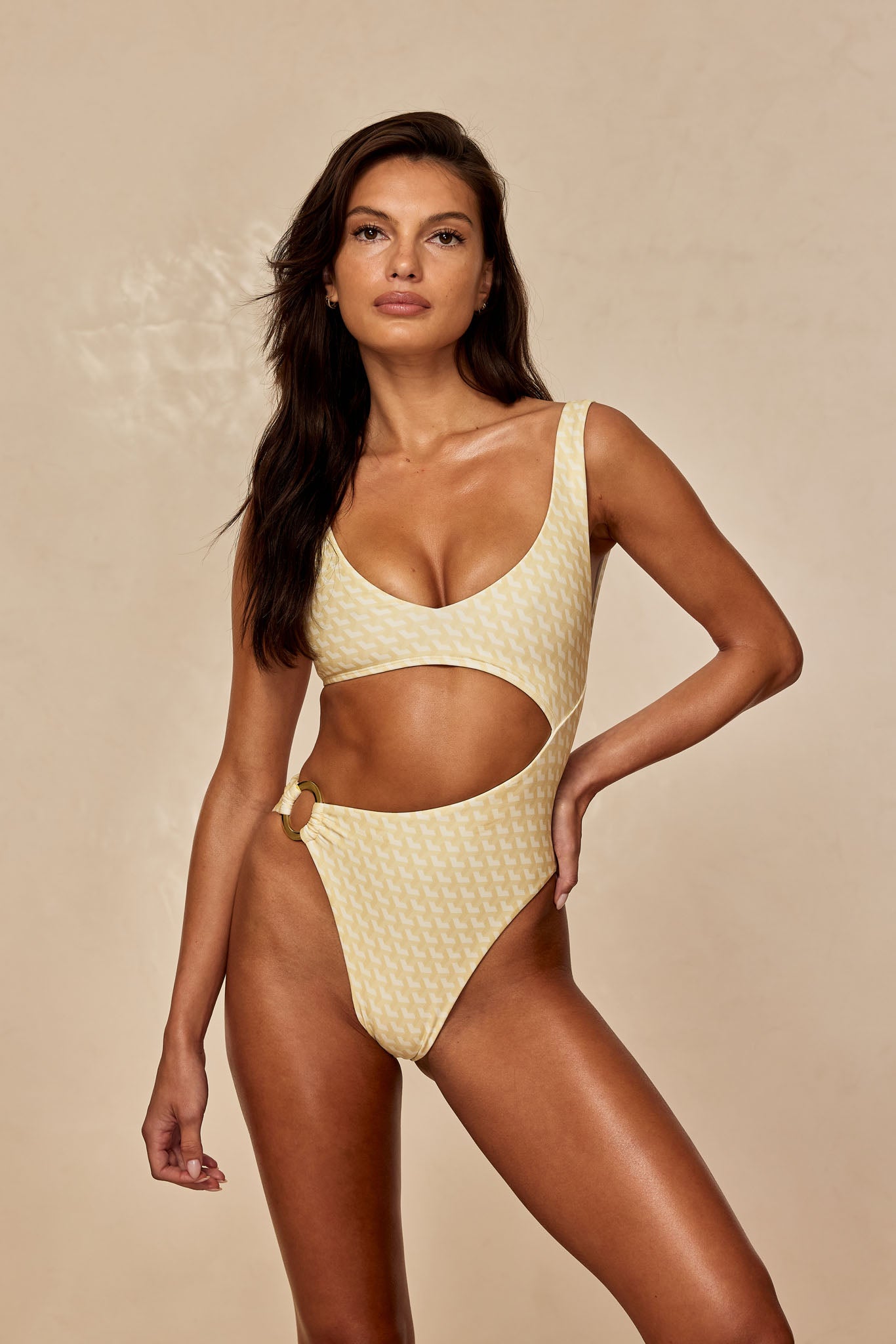 Model wearing yellow patterned one-piece swimsuit with side cutout and decorative ring on beige background