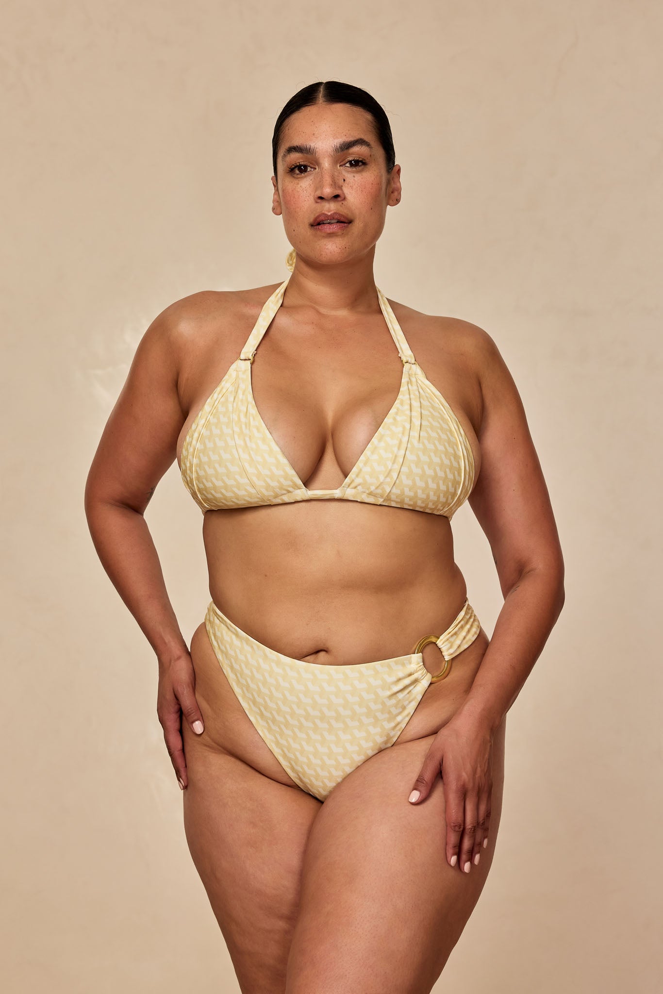 Model wearing a yellow patterned halter bikini with gold ring on high-cut bottoms, standing against beige backdrop.