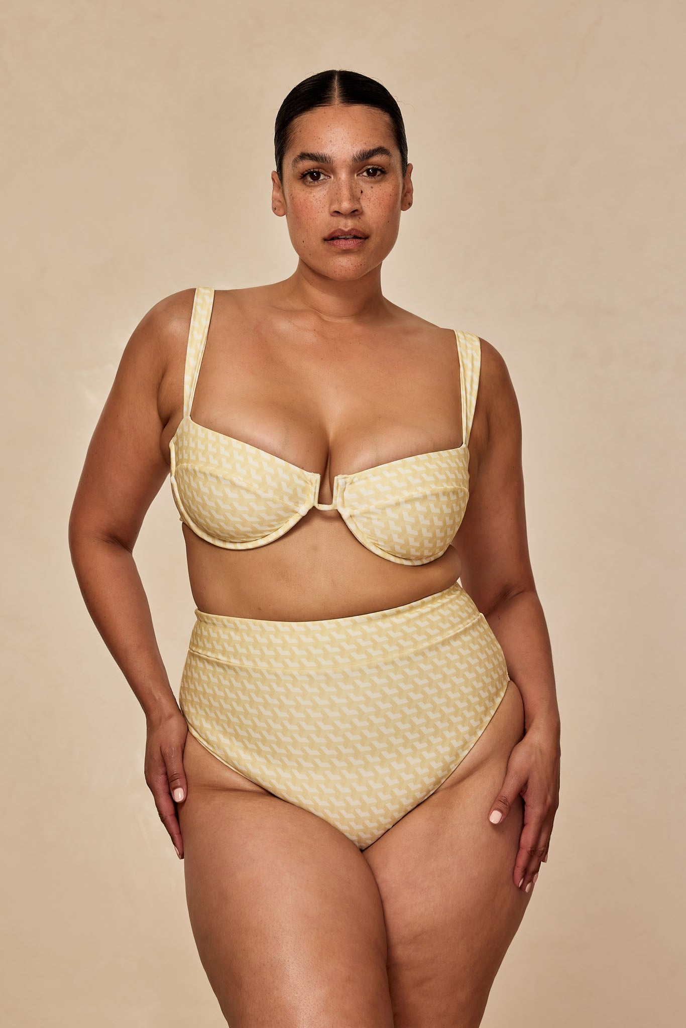 Plus-size model wearing yellow houndstooth-pattern underwire bikini top and high-waist bottoms on beige background