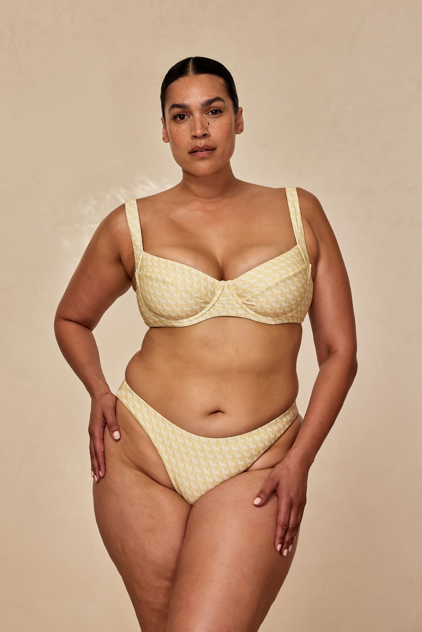 Woman wearing yellow patterned underwire bikini standing facing camera with hands on hips against beige background