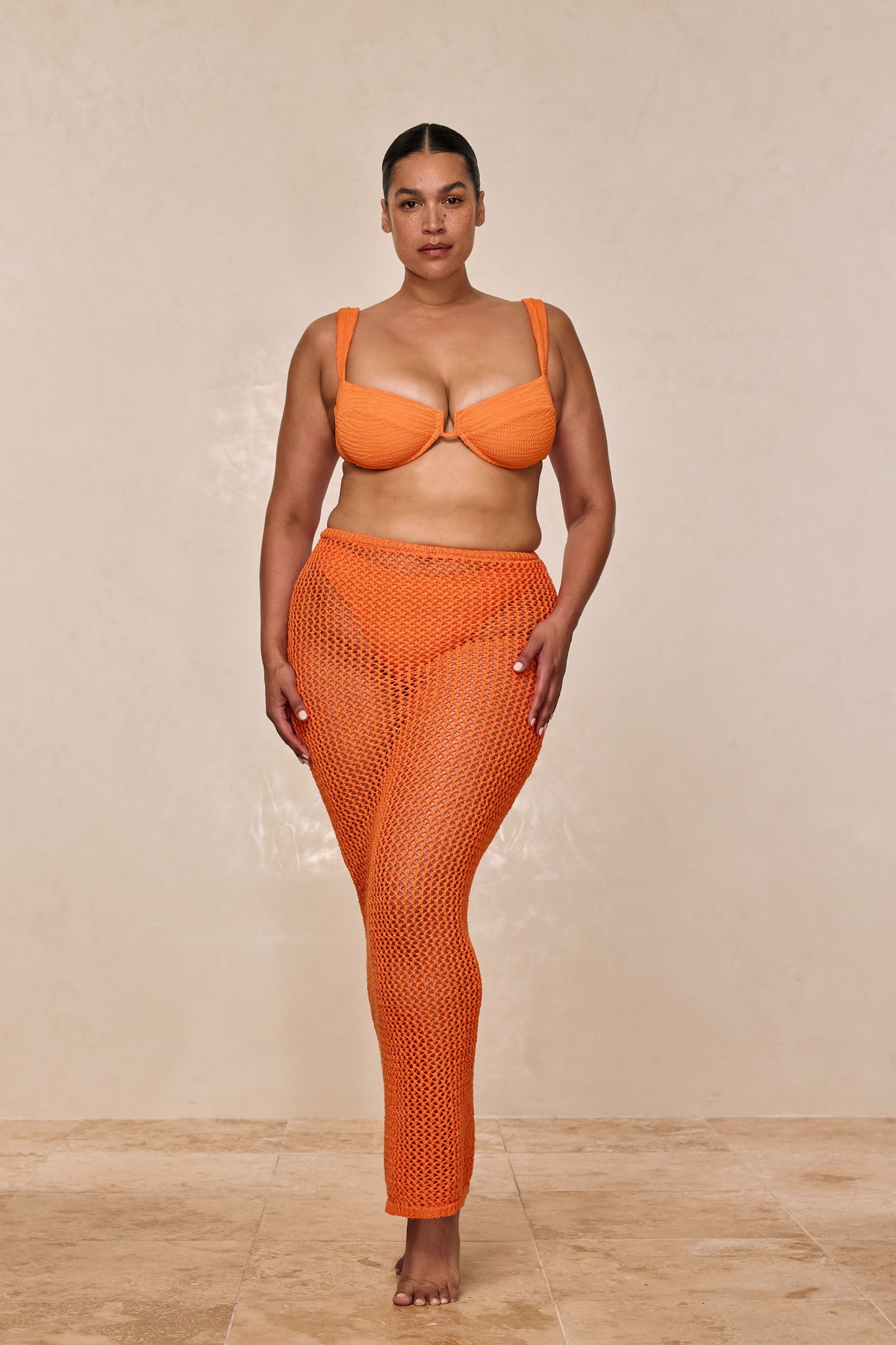 Plus-size woman standing barefoot in orange bikini top and crocheted mesh pants against a neutral background.