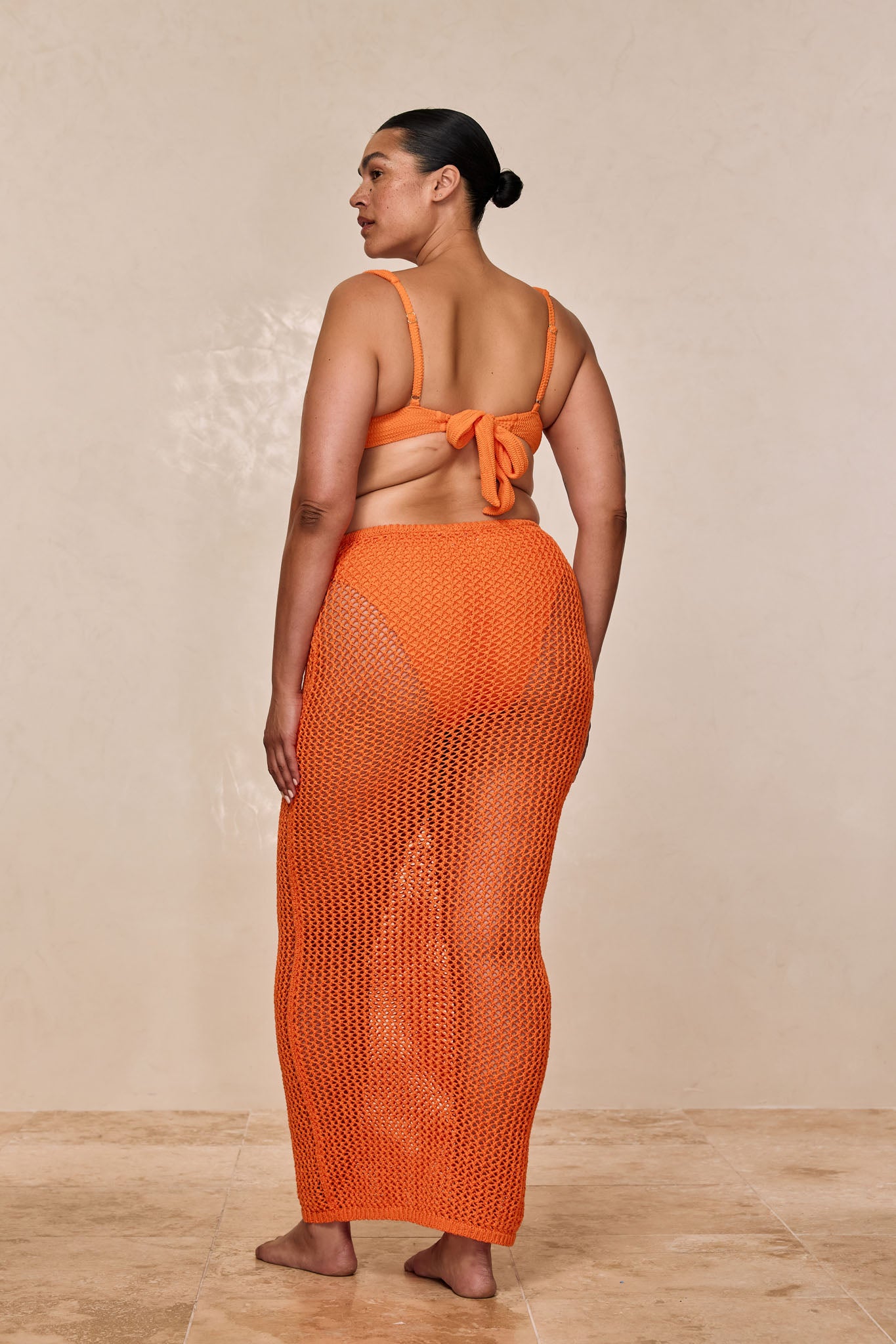 Back view of model wearing orange crocheted mesh maxi skirt over bikini bottoms with matching tie-back bikini top.