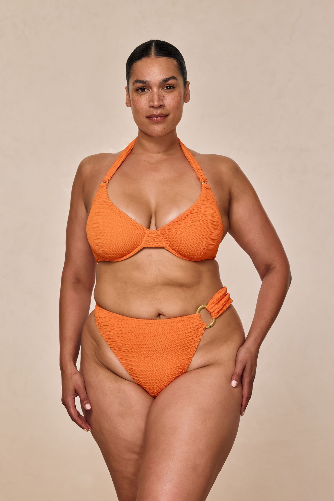 Woman modeling orange textured halter bikini with gold ring at hip, neutral studio background