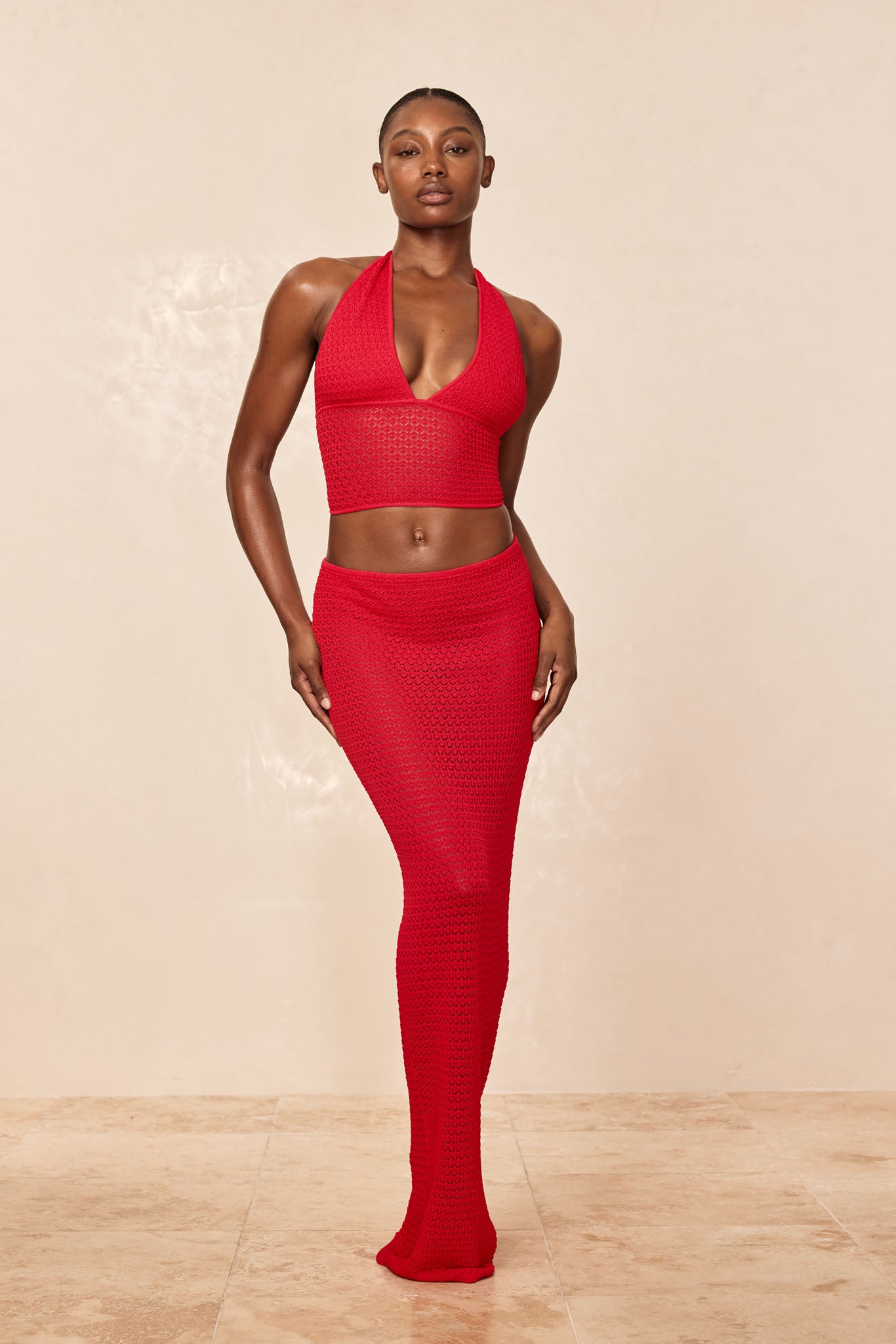 Woman wearing red crochet halter crop top and high-waisted long crochet skirt, standing against beige backdrop