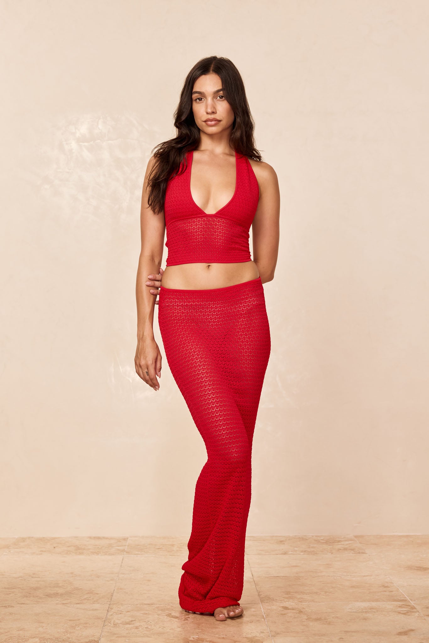 Woman modeling red crochet plunge halter crop top and matching long skirt, standing barefoot against beige studio background