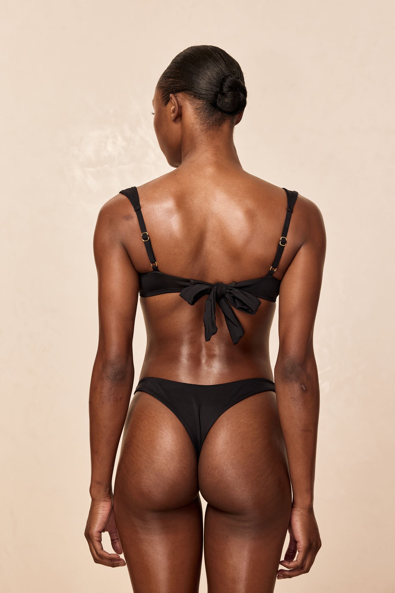 Back view of model wearing black tie-back bikini, low bun hairstyle, showing bow knot at center back.