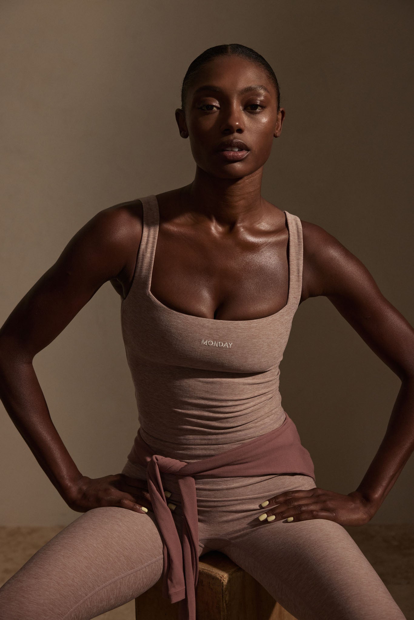 Seated athletic woman in taupe tank and leggings, hands on hips, 'MONDAY' logo on tank