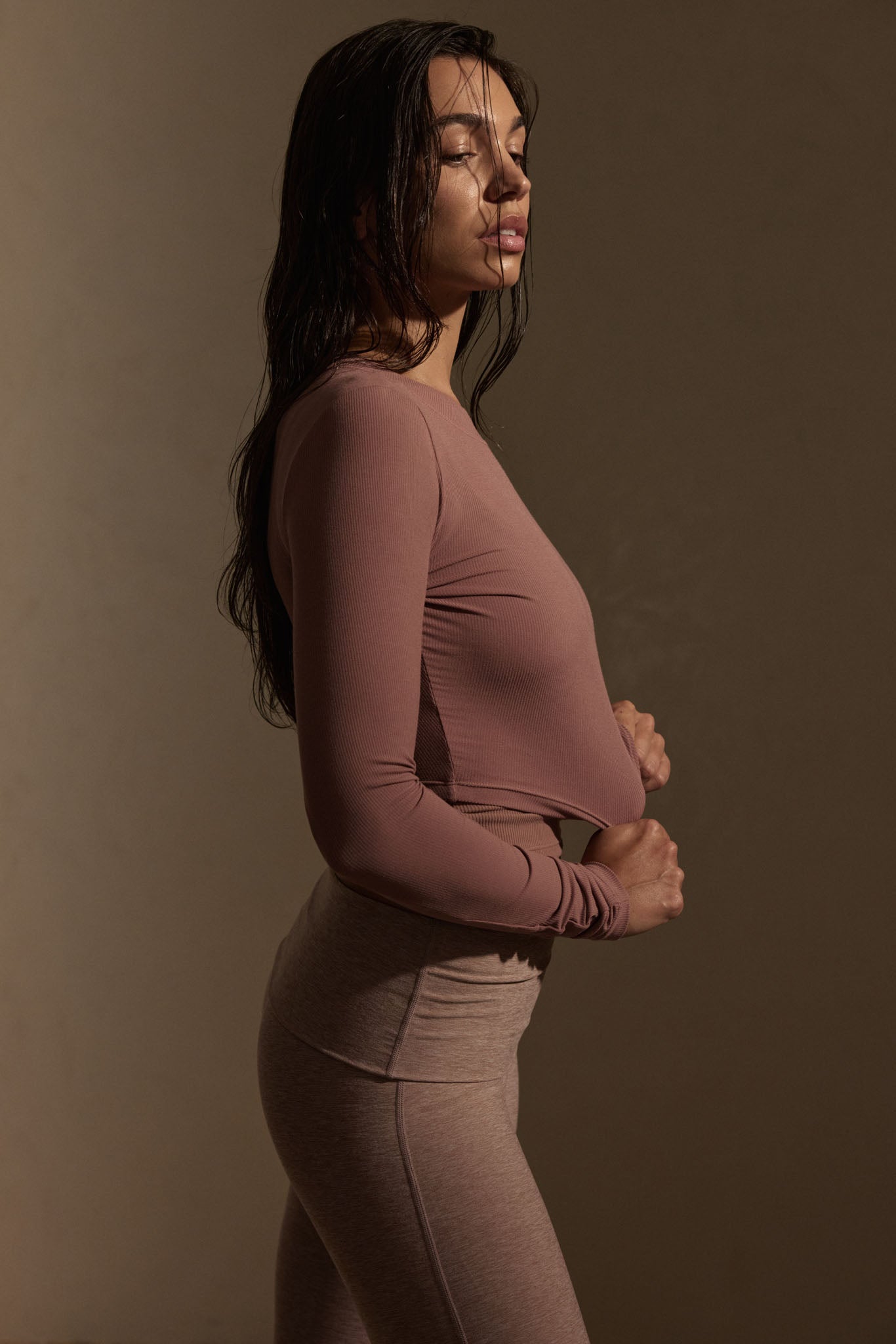 Side-profile of a woman with wet hair wearing a mauve long-sleeve top and matching high-waist leggings, gripping her top hem.