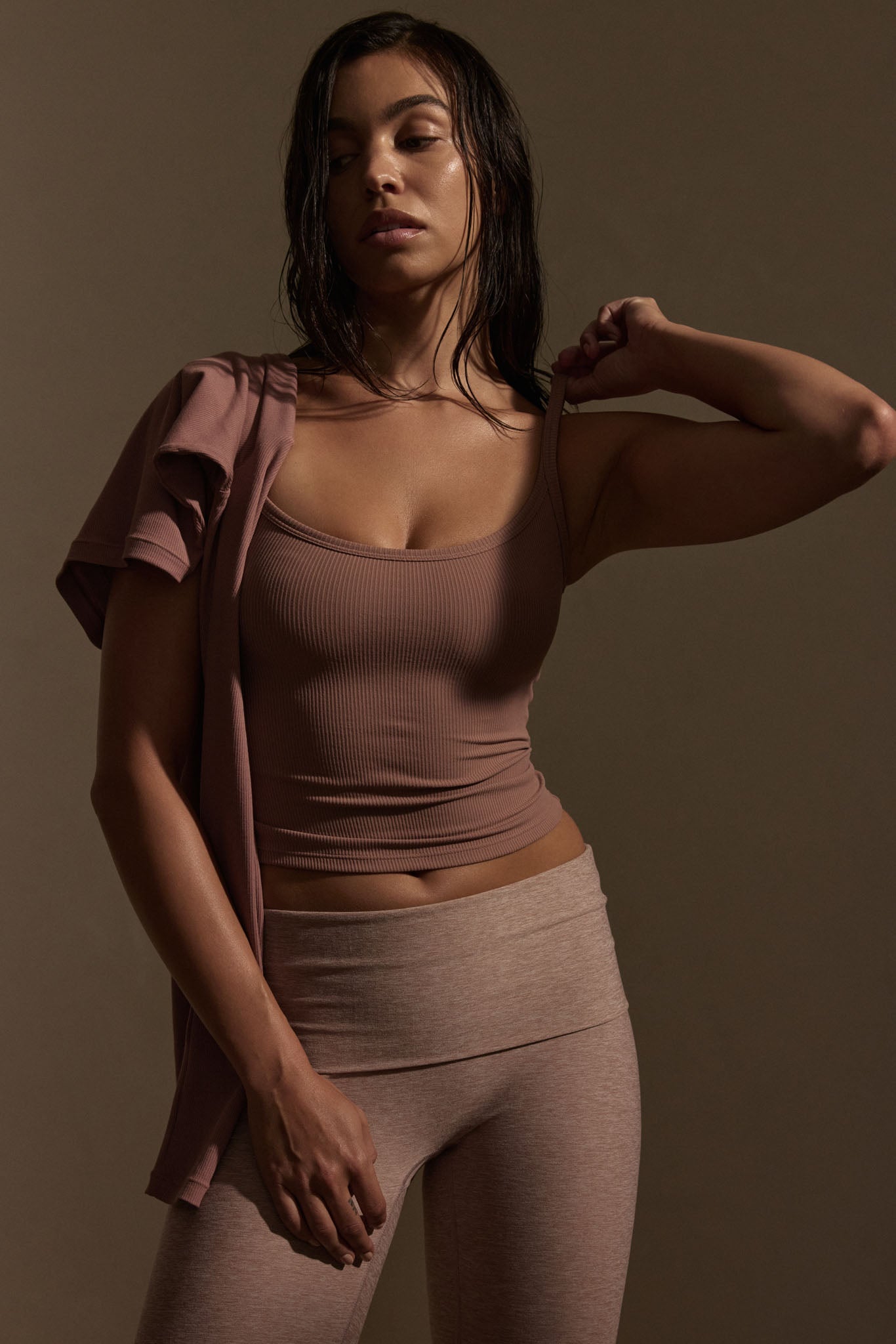 Woman in mauve ribbed tank and leggings adjusting strap, wet hair, studio-lit neutral background