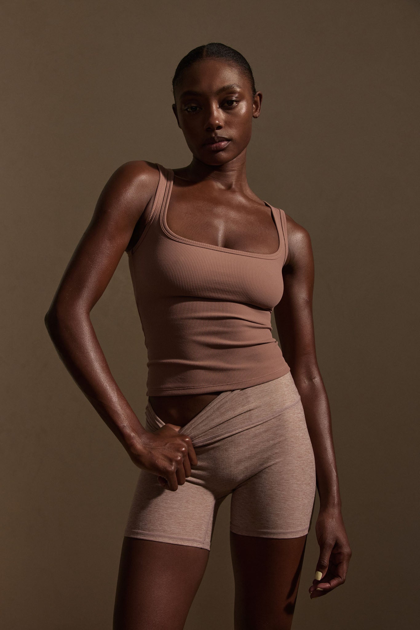 Model wearing a brown ribbed tank and beige biker shorts, pulling her waistband in a studio portrait.