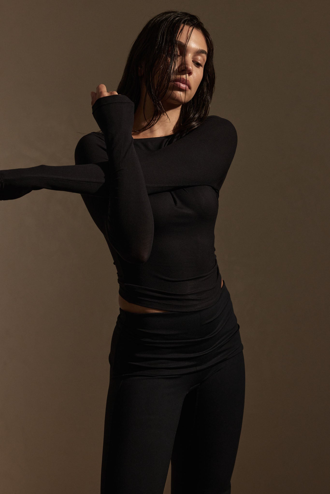 Woman with wet hair stretching arms while wearing a fitted black long-sleeve top and black leggings against a brown backdrop.