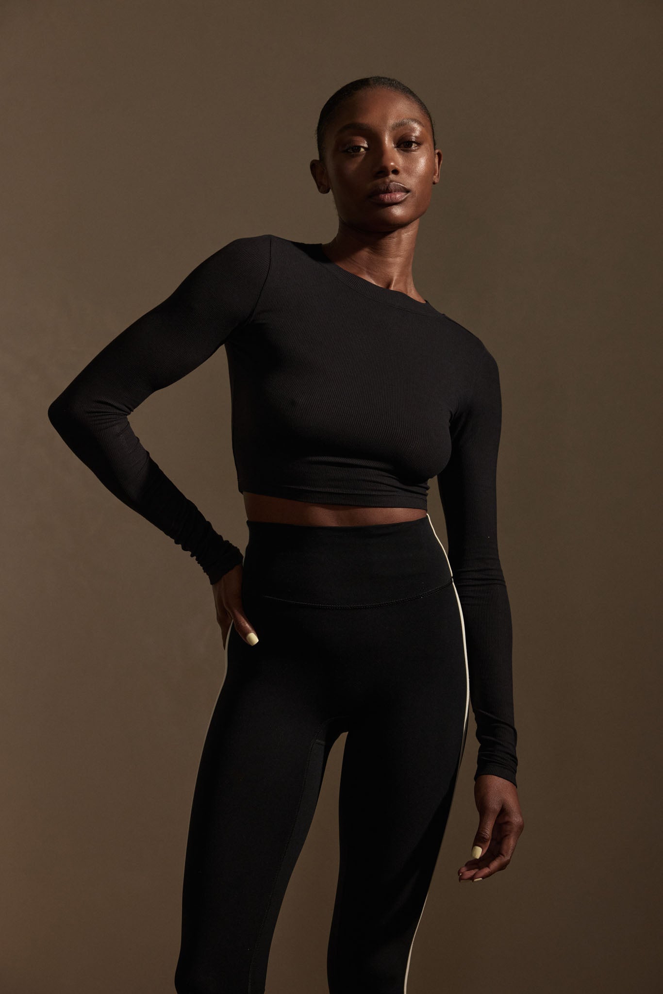 Woman wearing black cropped long-sleeve top and high-waist leggings with side stripe, standing against brown background