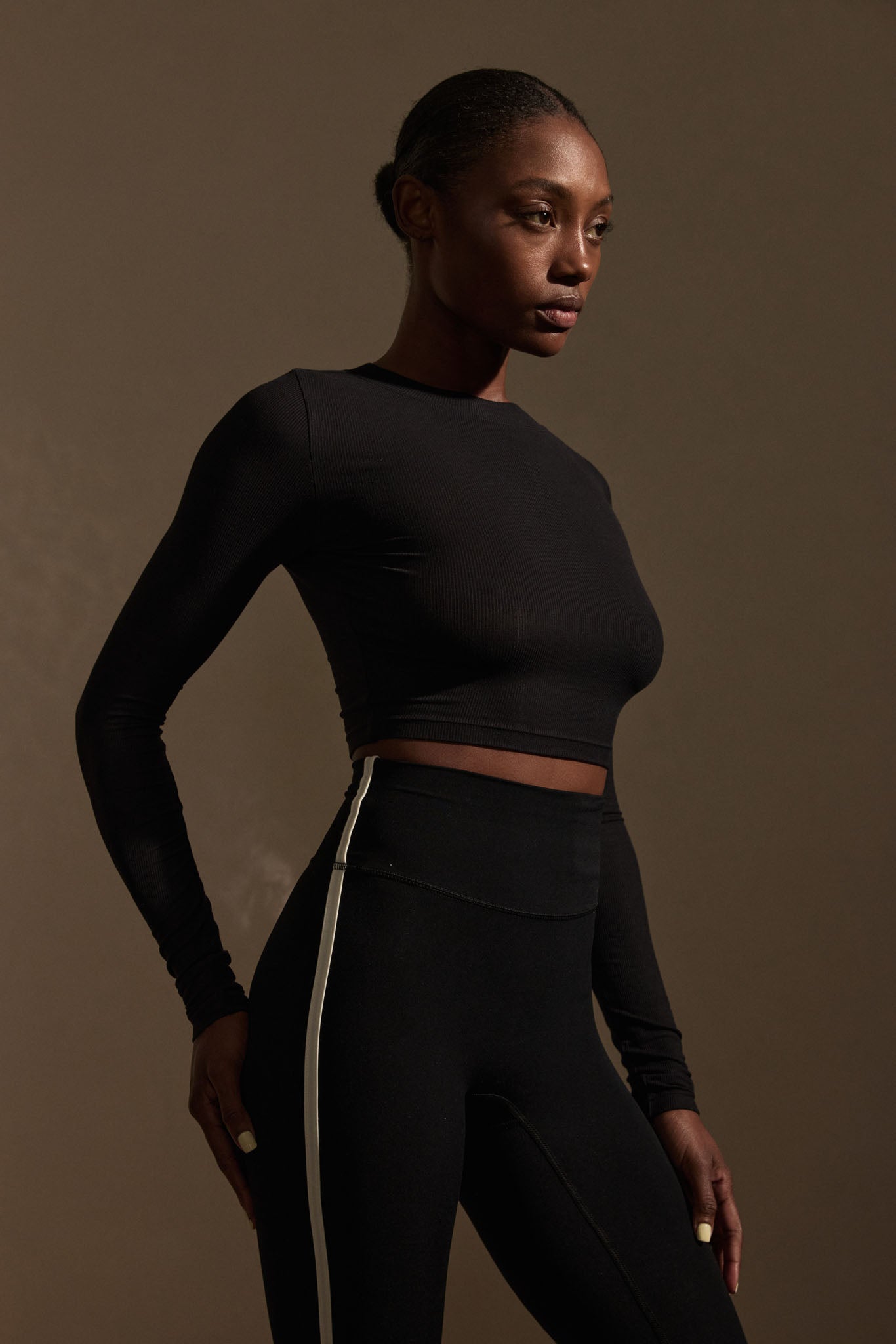 Woman in black long-sleeve crop top and high-waist leggings with white side stripe, three-quarter pose looking right.