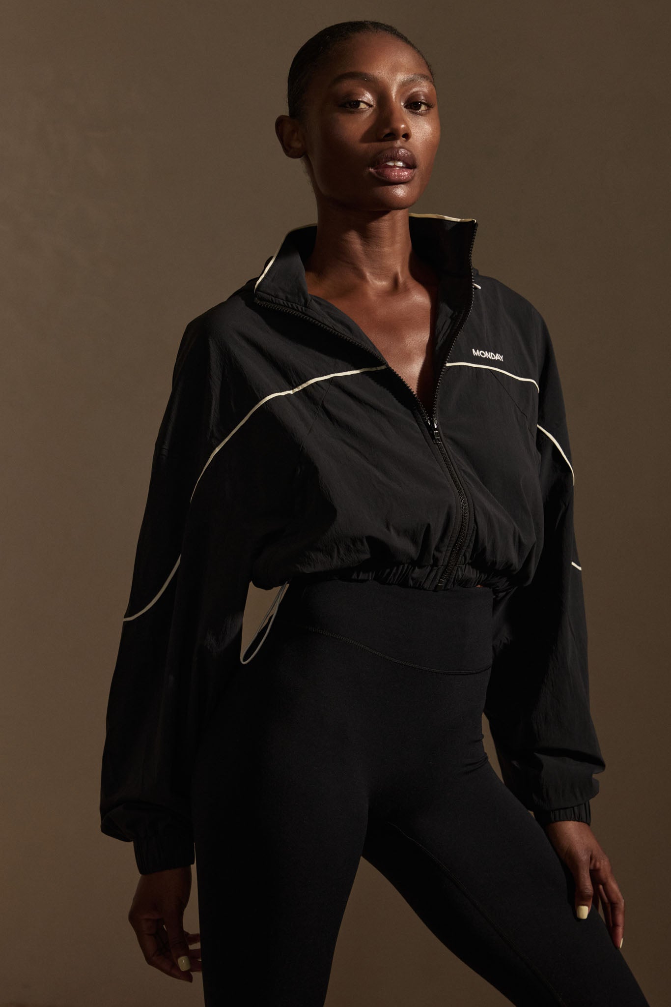 Female model wearing a black cropped zip jacket with 'MONDAY' logo and high-waist black leggings, neutral background