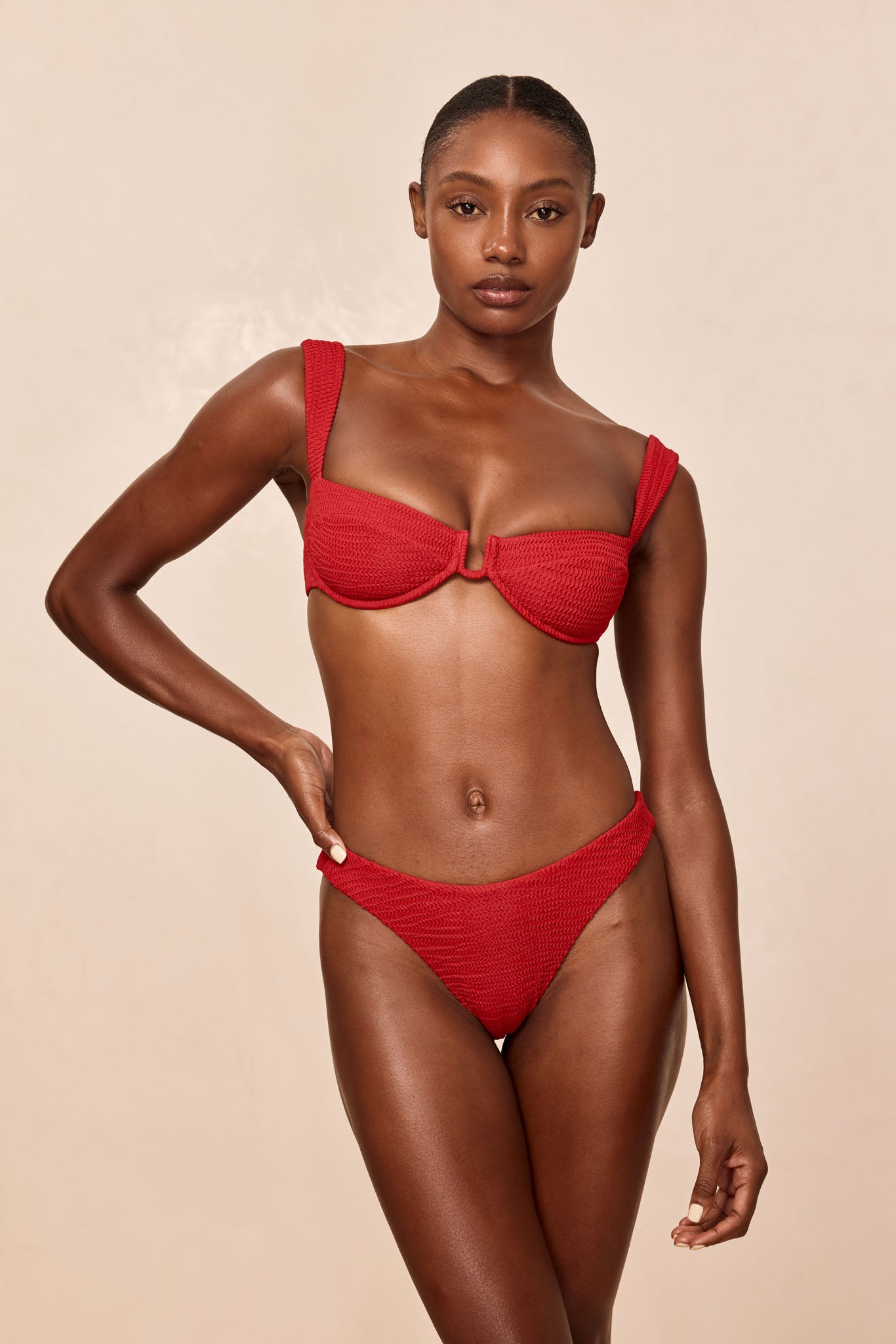 Woman modeling red textured bikini, front view, standing with one hand on hip against a neutral beige background.