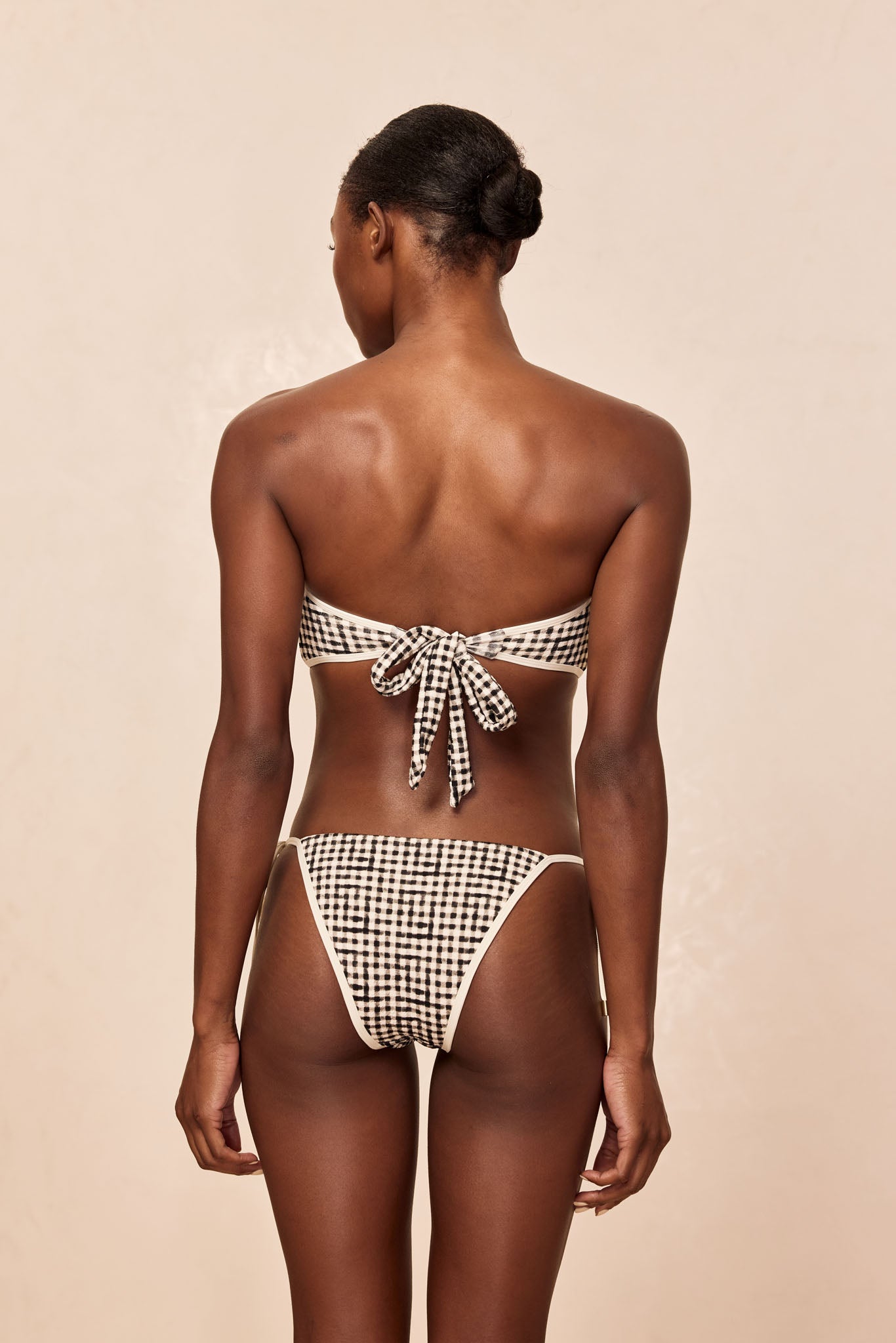 Rear view of woman wearing black-and-white gingham tie-back bandeau bikini, hair in a low bun, neutral background.