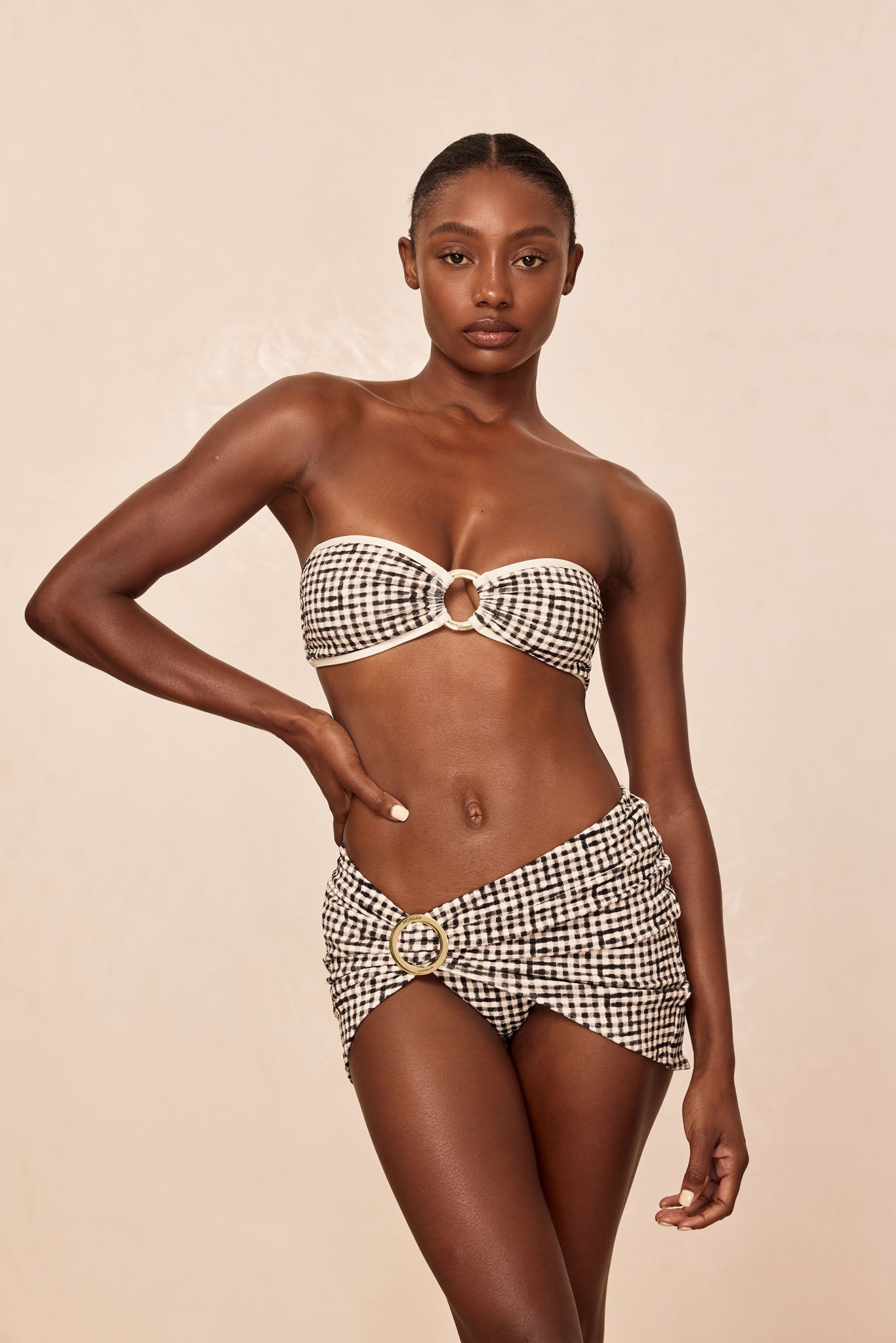 Model wearing black-and-white gingham bandeau bikini with gold ring detail and matching ruched sarong, hand on hip.