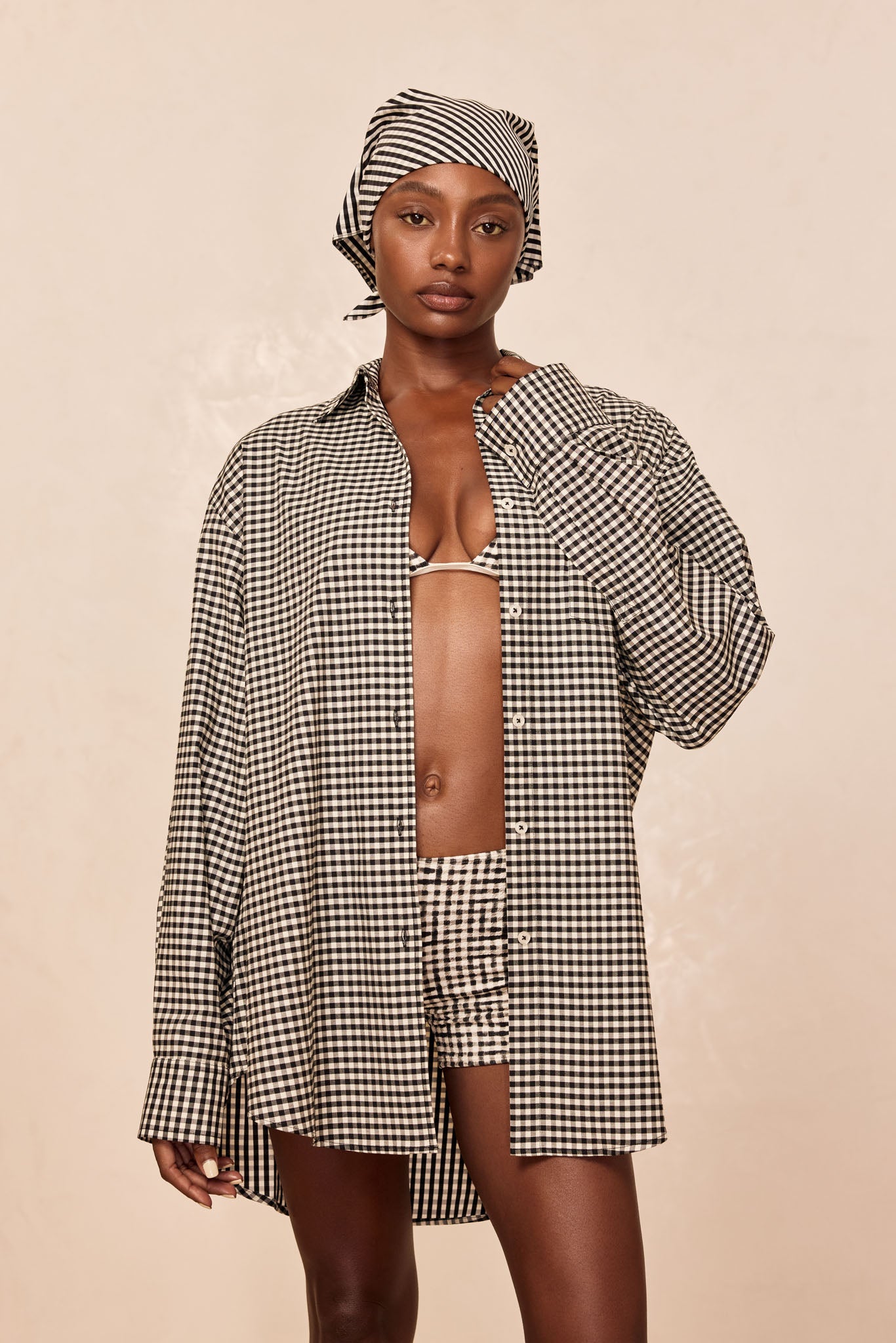 Woman wearing black-and-white gingham button shirt and headscarf, bikini top and shorts, posed against a neutral backdrop.