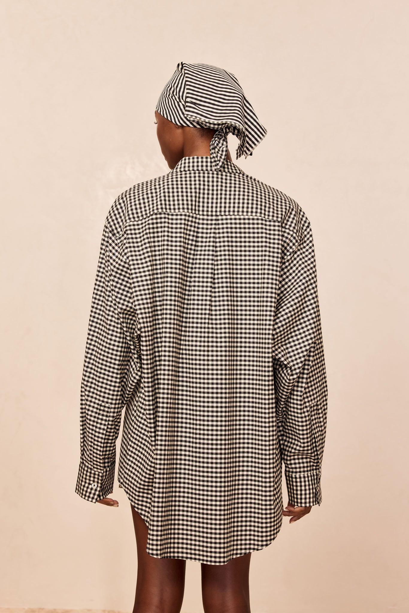 Back view of model wearing black-and-white gingham oversized shirt dress and matching headscarf against plain beige background