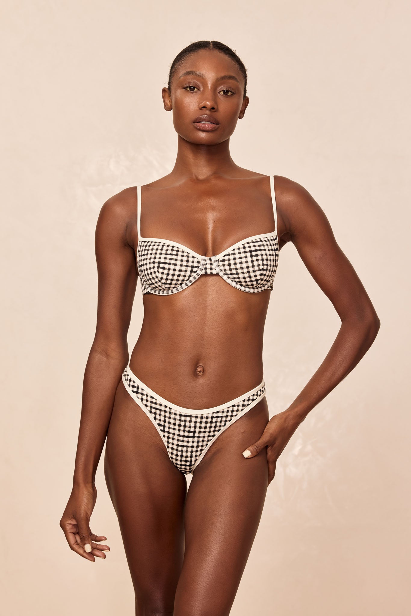 Female model wearing a black-and-white gingham underwire bra and matching high-cut briefs against a neutral beige background.