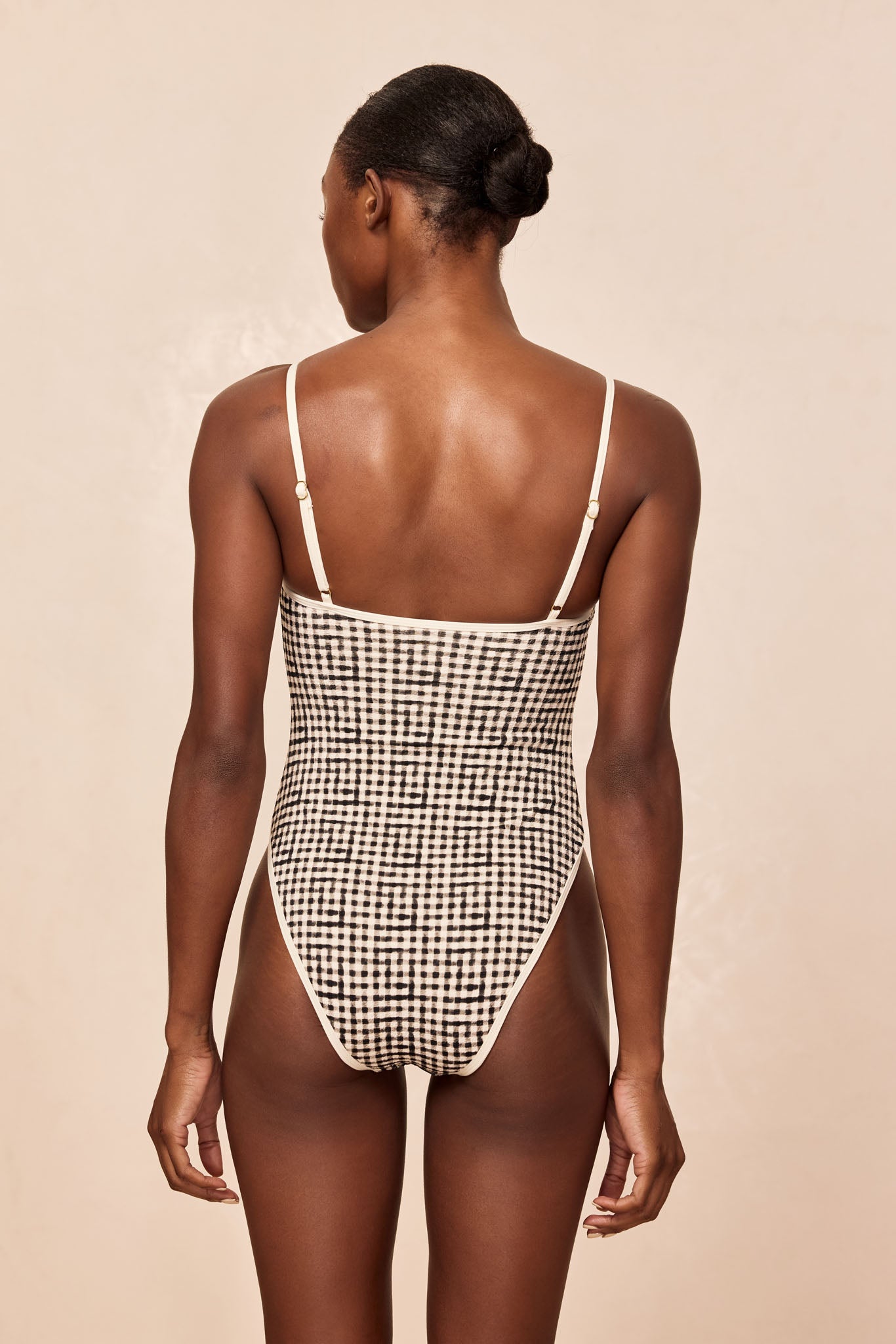 Back view of woman wearing high-cut black-and-white checkered one-piece swimsuit with thin adjustable cream straps