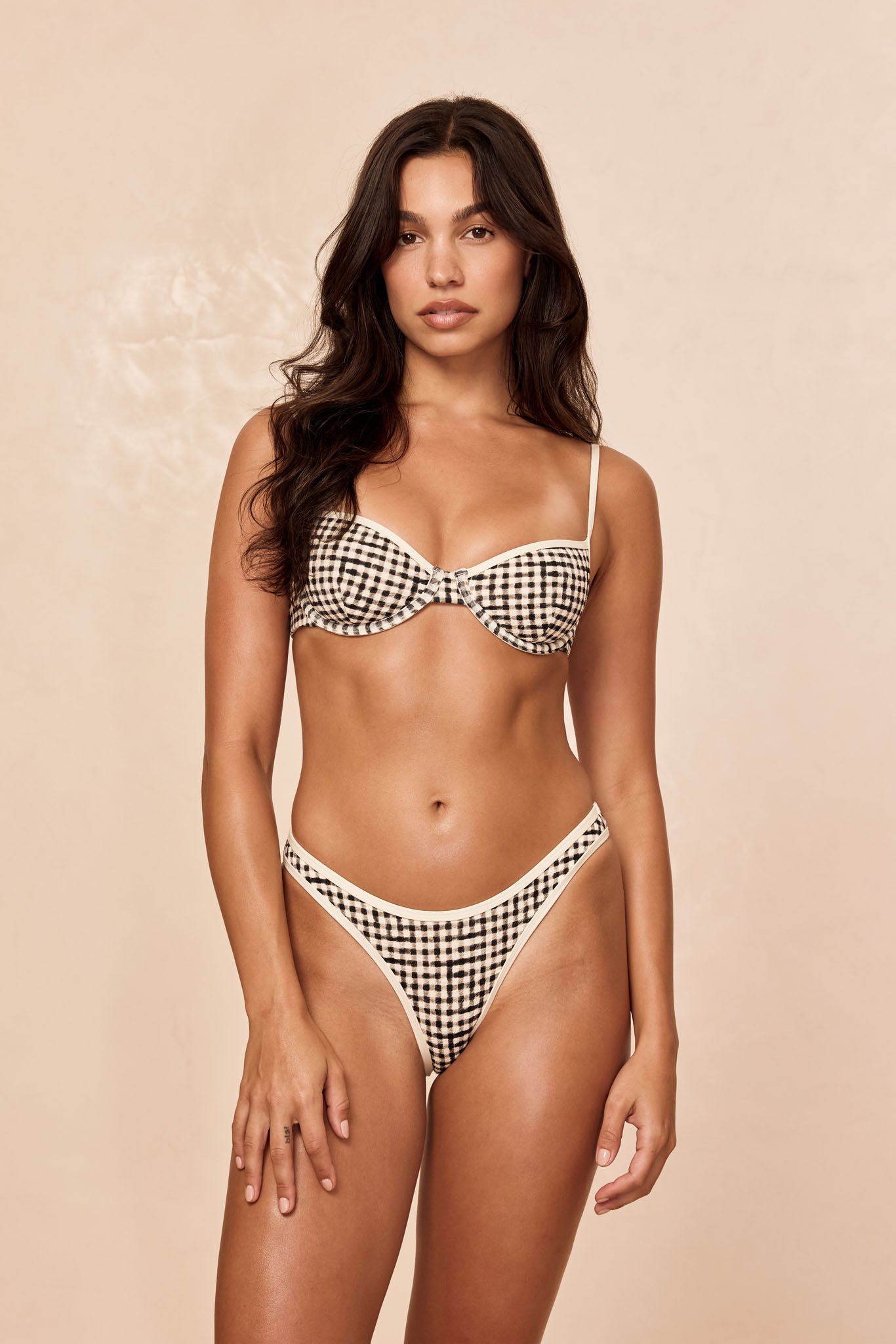Model wearing a black-and-cream gingham underwire bikini set, standing front-facing against a beige background.