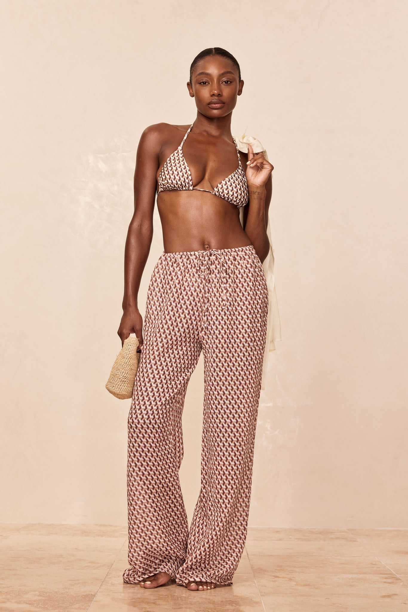 Woman wearing patterned triangle bikini top and matching wide-leg drawstring pants, holding woven clutch and cream cover-up.