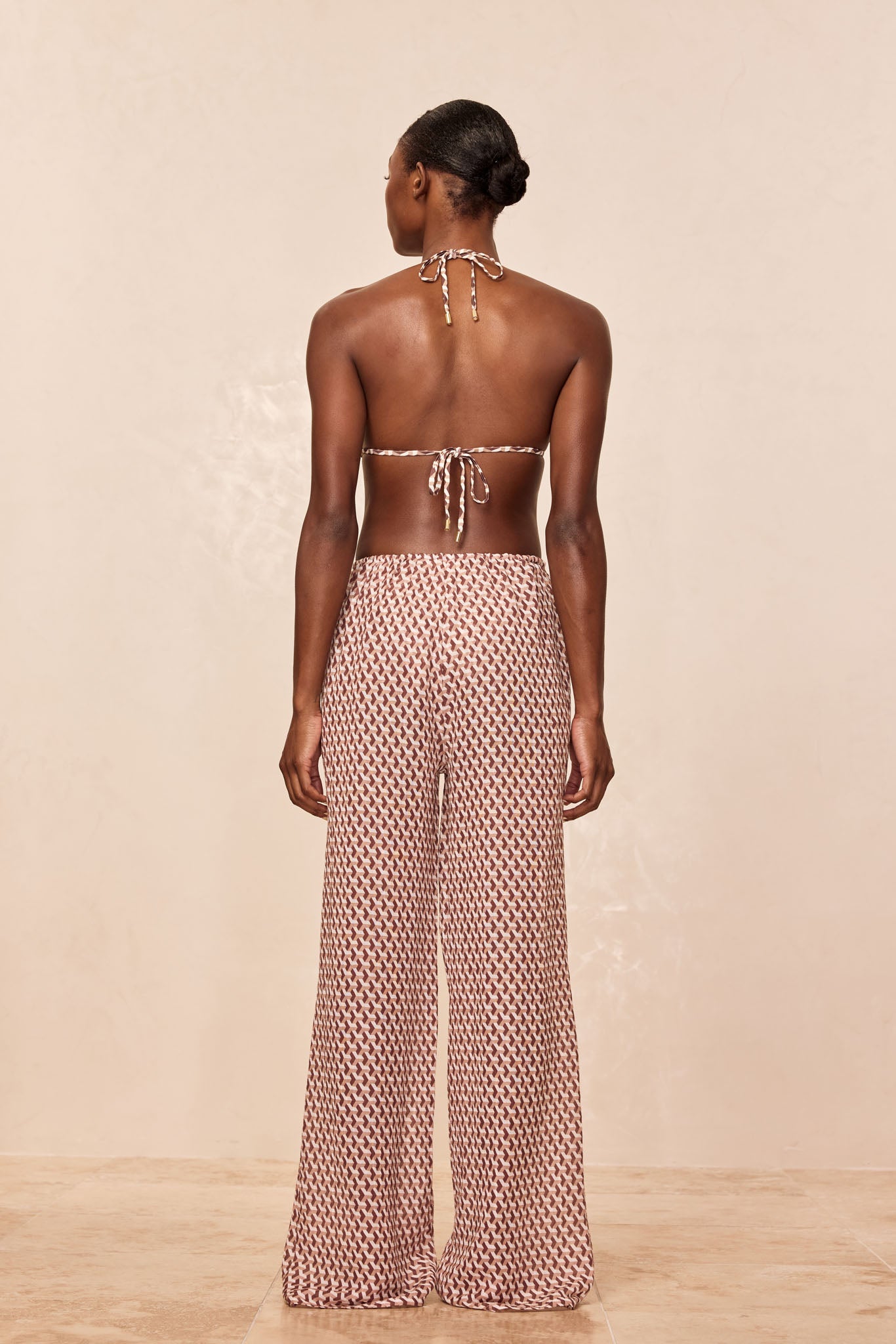 Back view of a woman in a tied halter bikini top and high-waist wide-leg patterned pants on a beige backdrop.