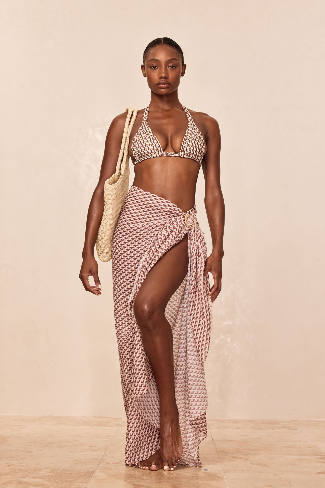 Model wearing brown geometric-print bikini top and matching sarong tied with a ring, carrying a woven beach tote