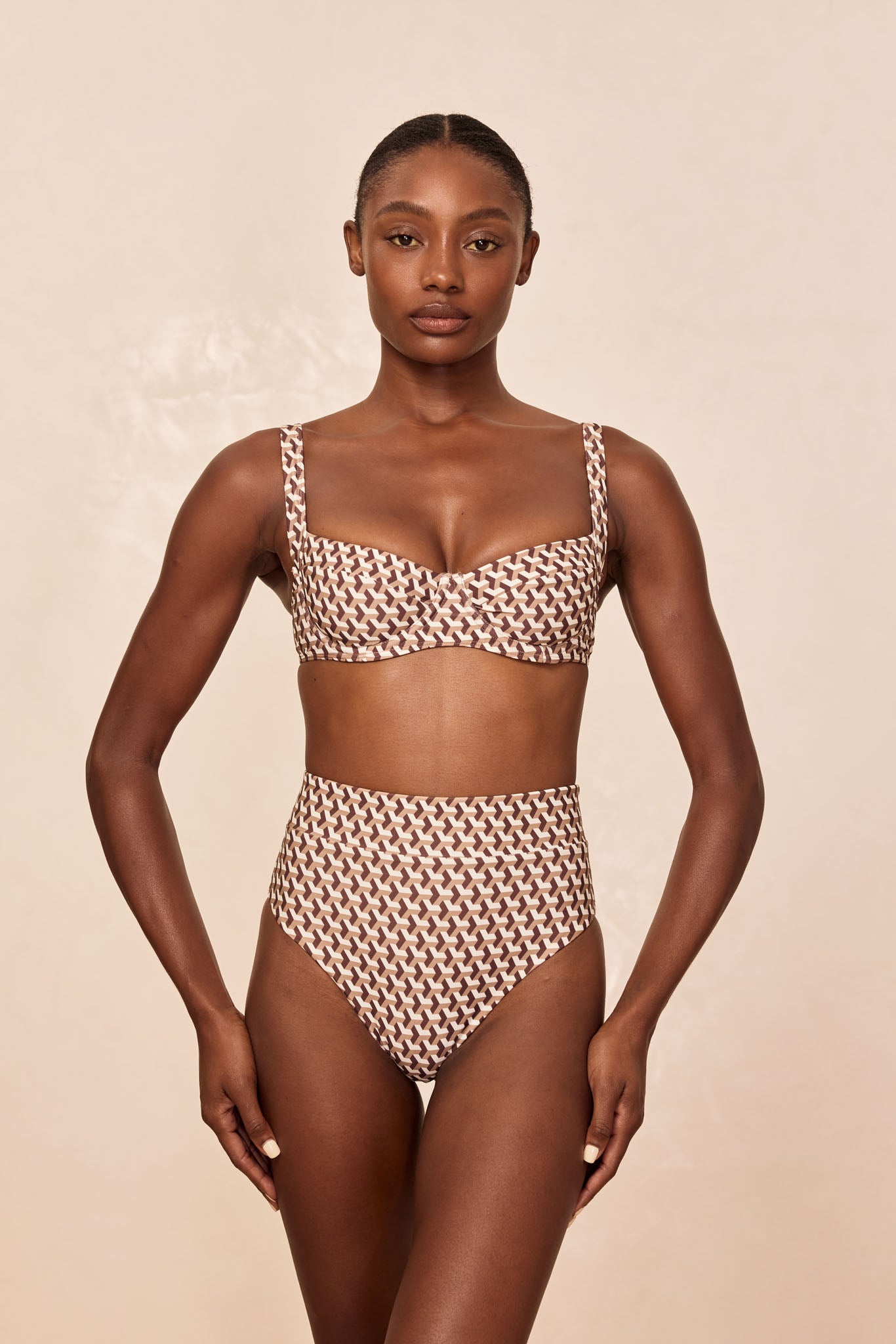 Model wearing brown-and-cream geometric high-waist bikini, front-facing pose on neutral beige background.