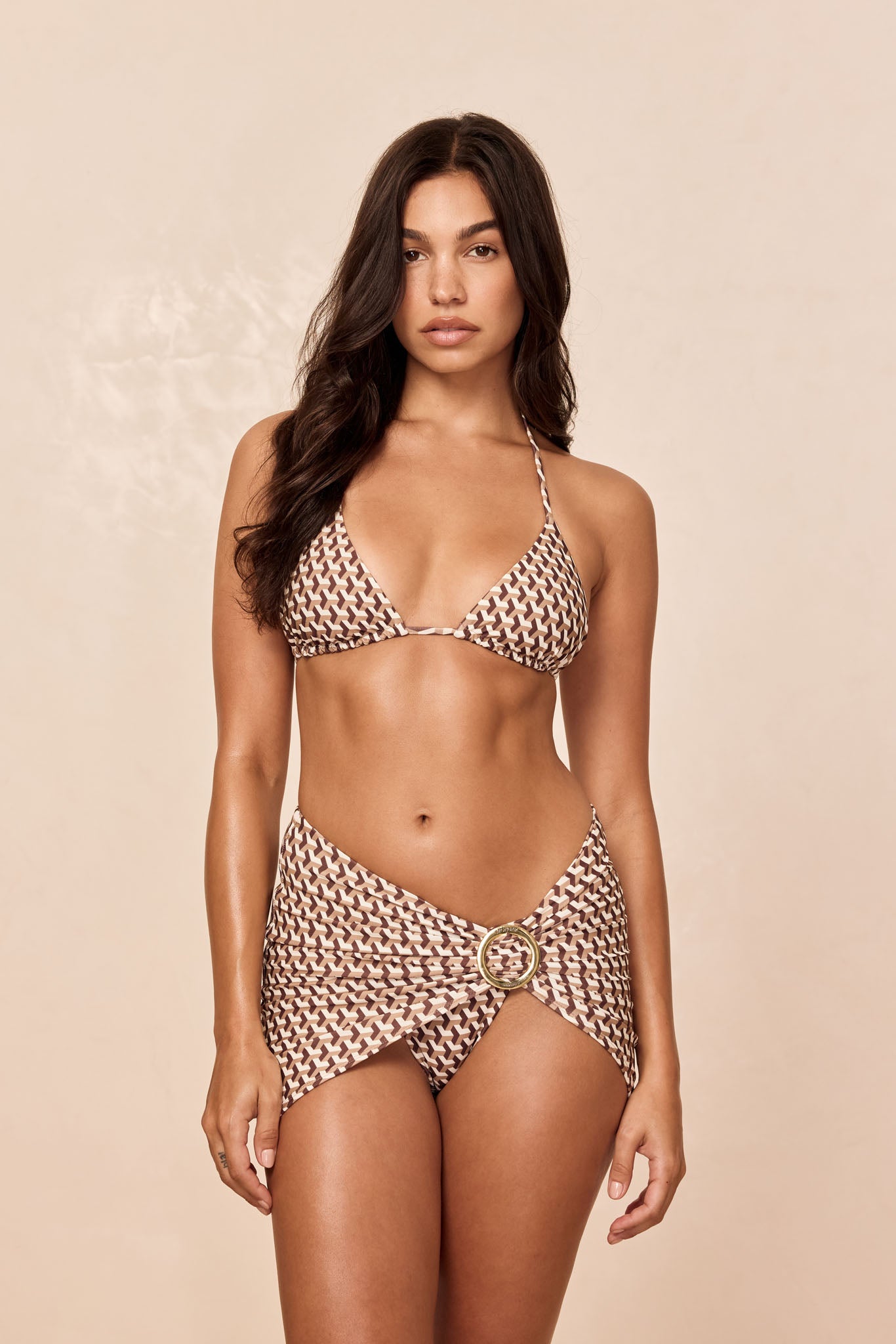 Woman modeling brown patterned triangle bikini top and matching sarong skirt with gold ring at hip, neutral background.
