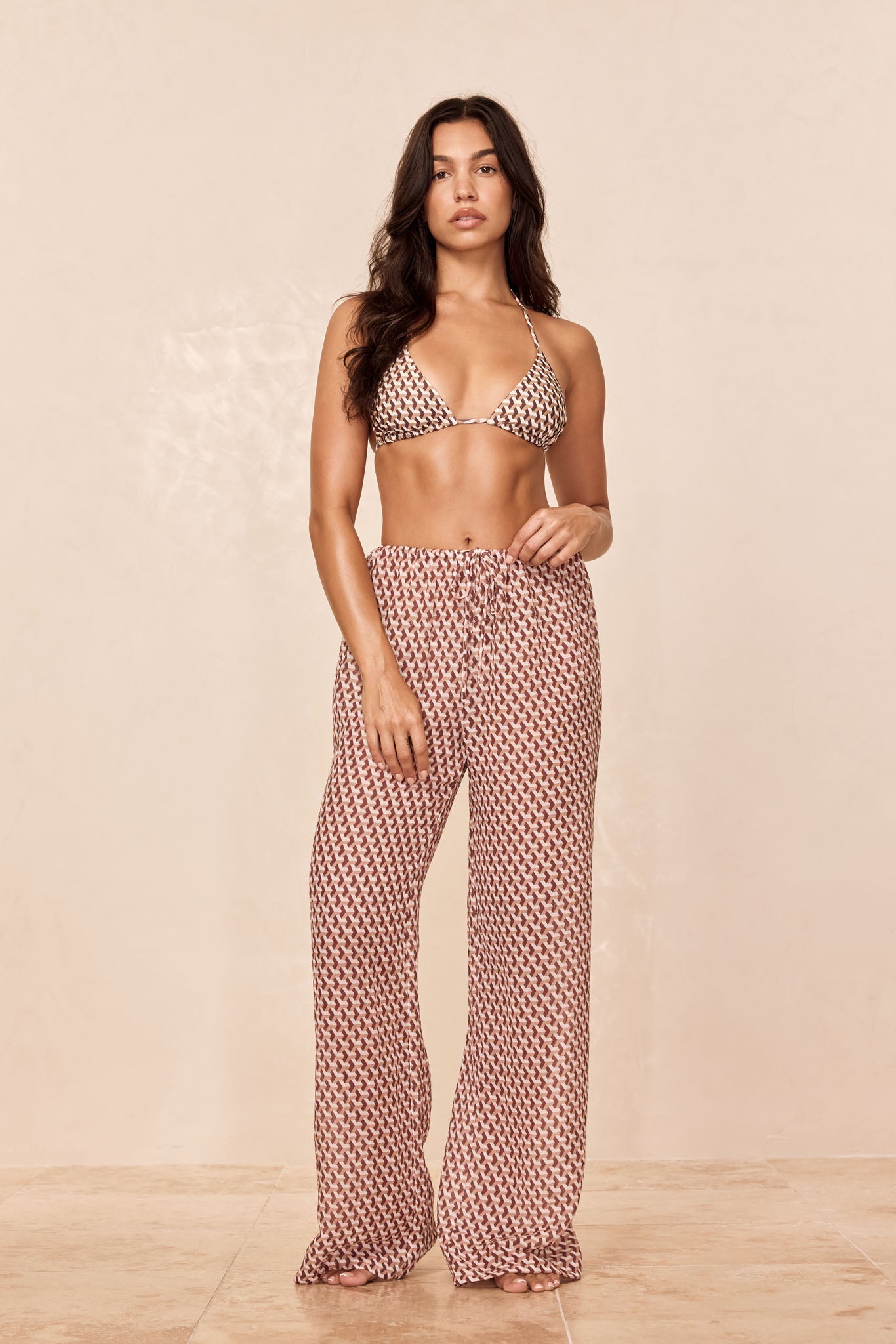 Woman wearing a triangle bikini top and drawstring wide-leg patterned pants on a beige studio background.