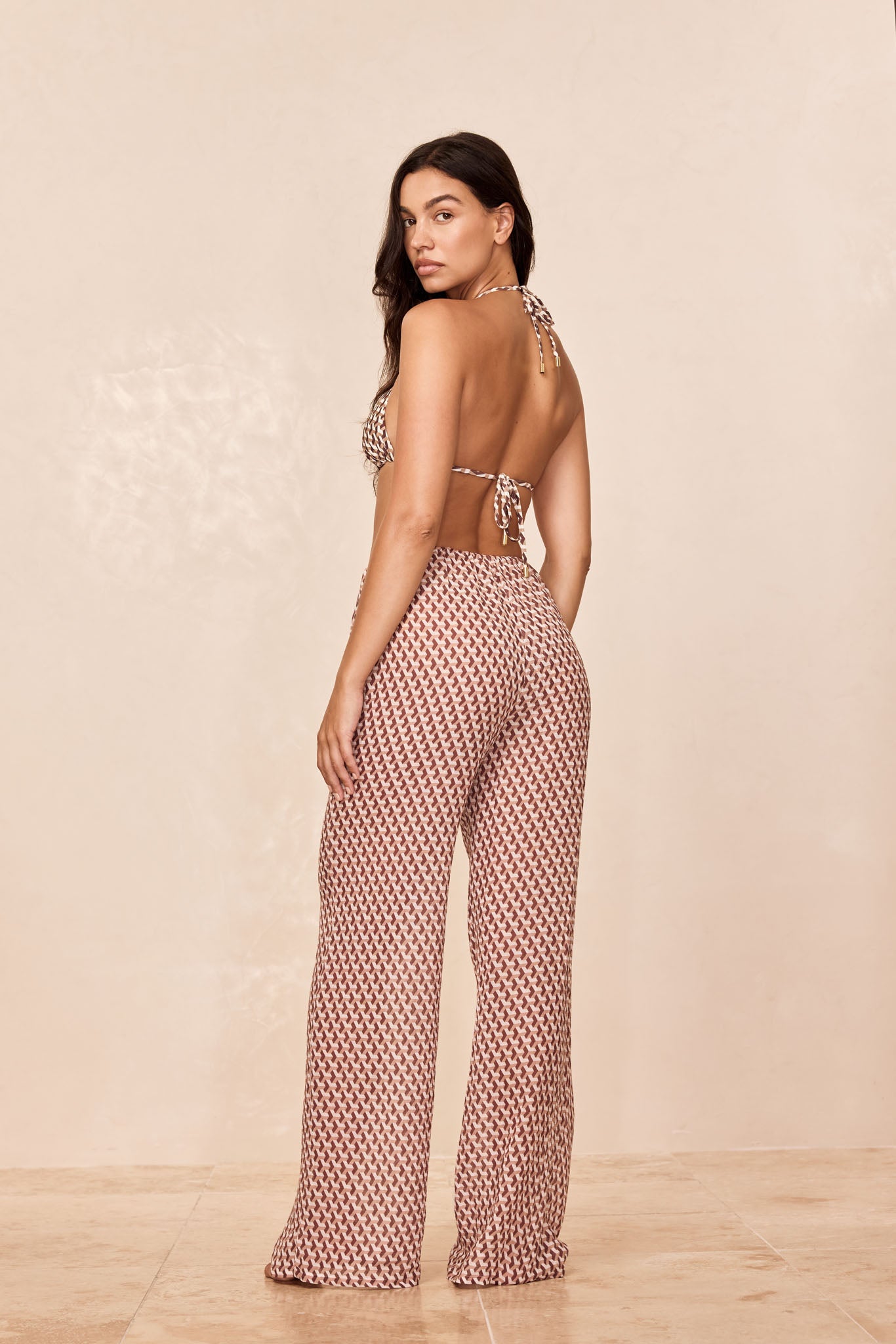 Model standing with back to camera, looking over her shoulder, wearing halter bikini top and high-waist patterned wide-leg pants.