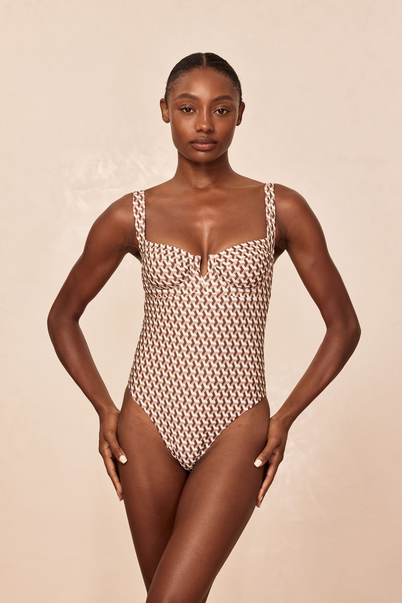 Female model facing camera wearing brown-and-cream geometric one-piece swimsuit on neutral beige background.