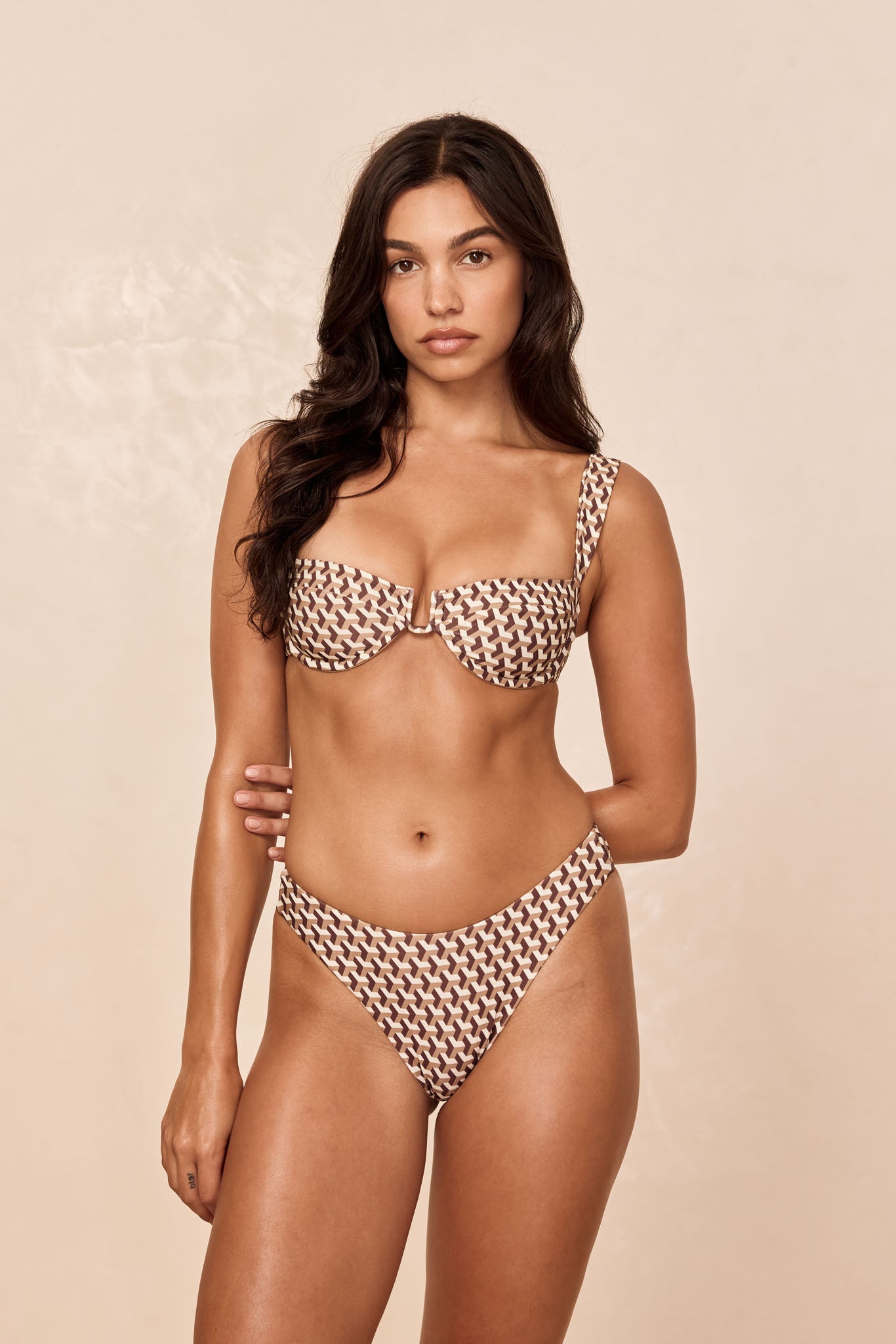 Woman wearing brown and cream geometric-print bikini with underwire top and high-cut bottoms, against a neutral beige backdrop.