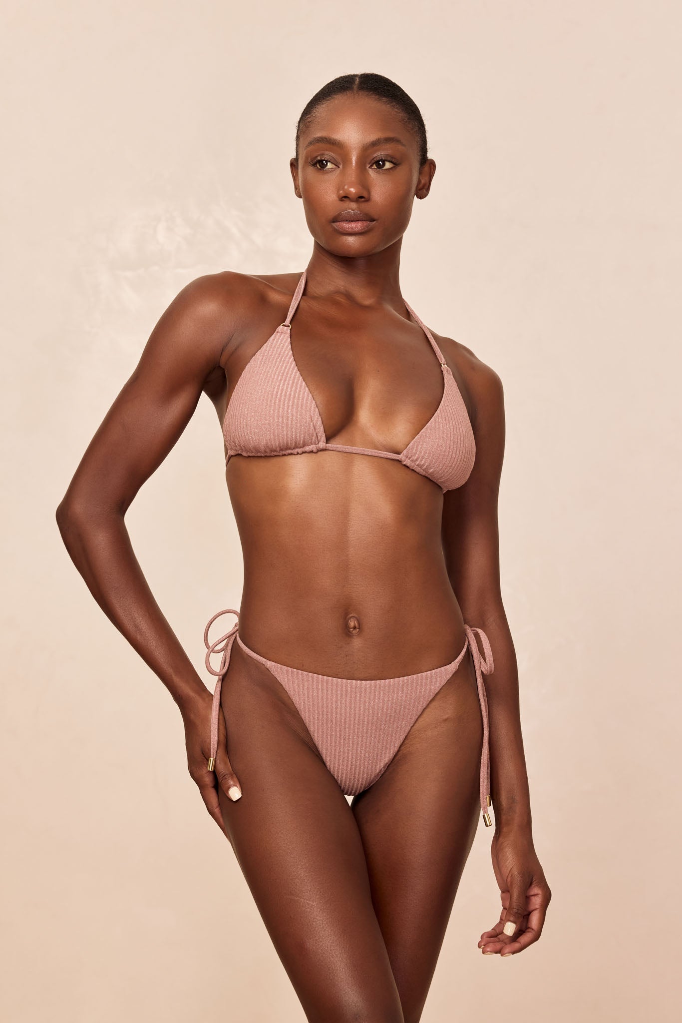 Model wearing mauve ribbed triangle bikini with halter straps and side-tie bottoms, standing against pale beige background.