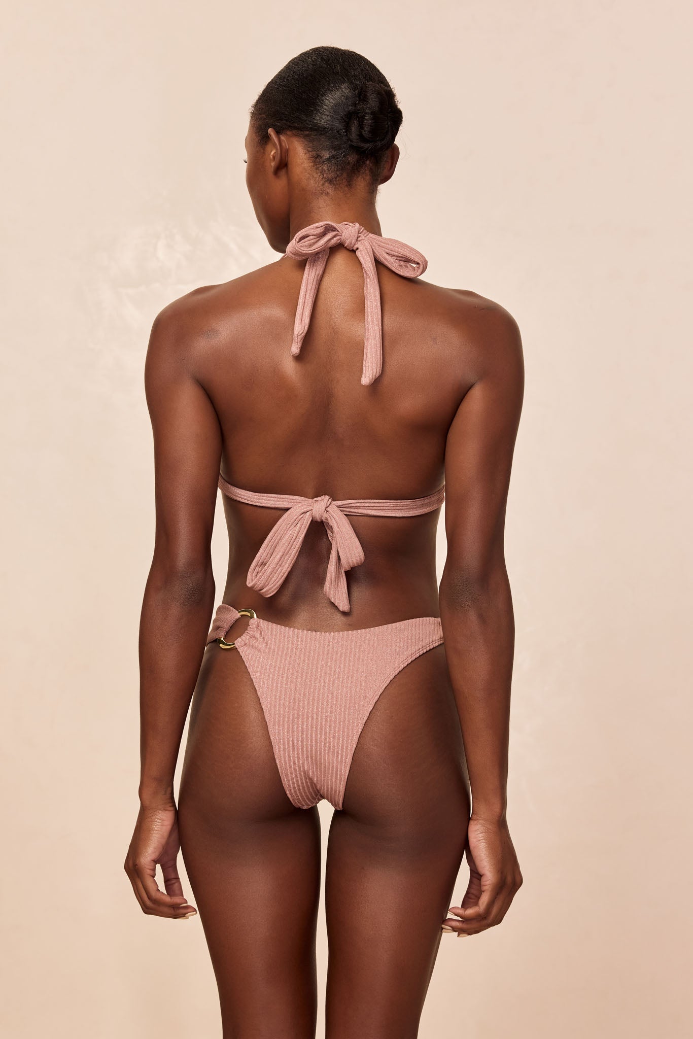 Back view of model wearing mauve ribbed halter bikini, tied bows at neck and back, gold ring on left hip