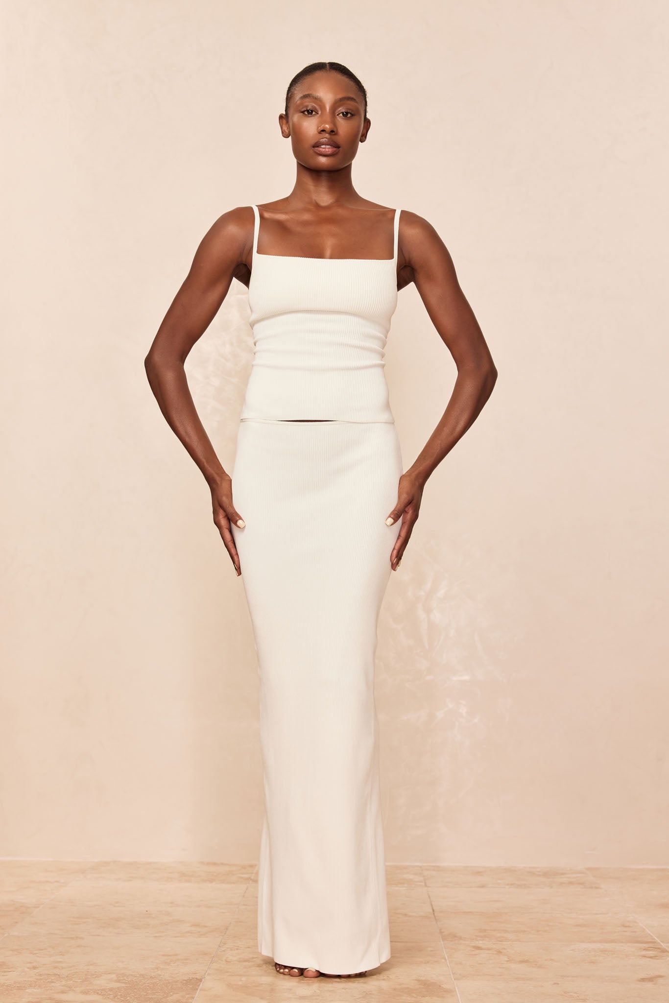 Model facing camera in a cream ribbed square-neck tank and matching long fitted maxi skirt on neutral background.