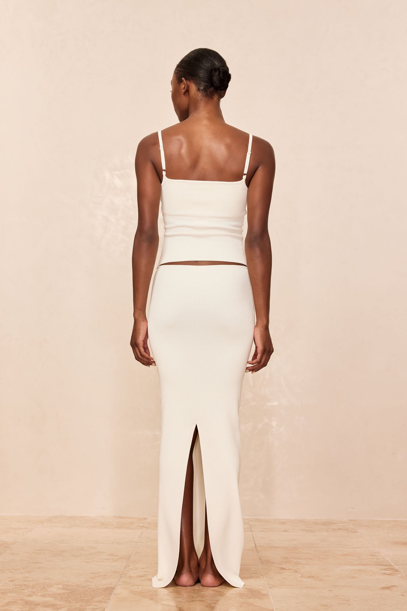 Back view of woman wearing cream ribbed camisole and matching long skirt with center back slit, hair in a low bun.