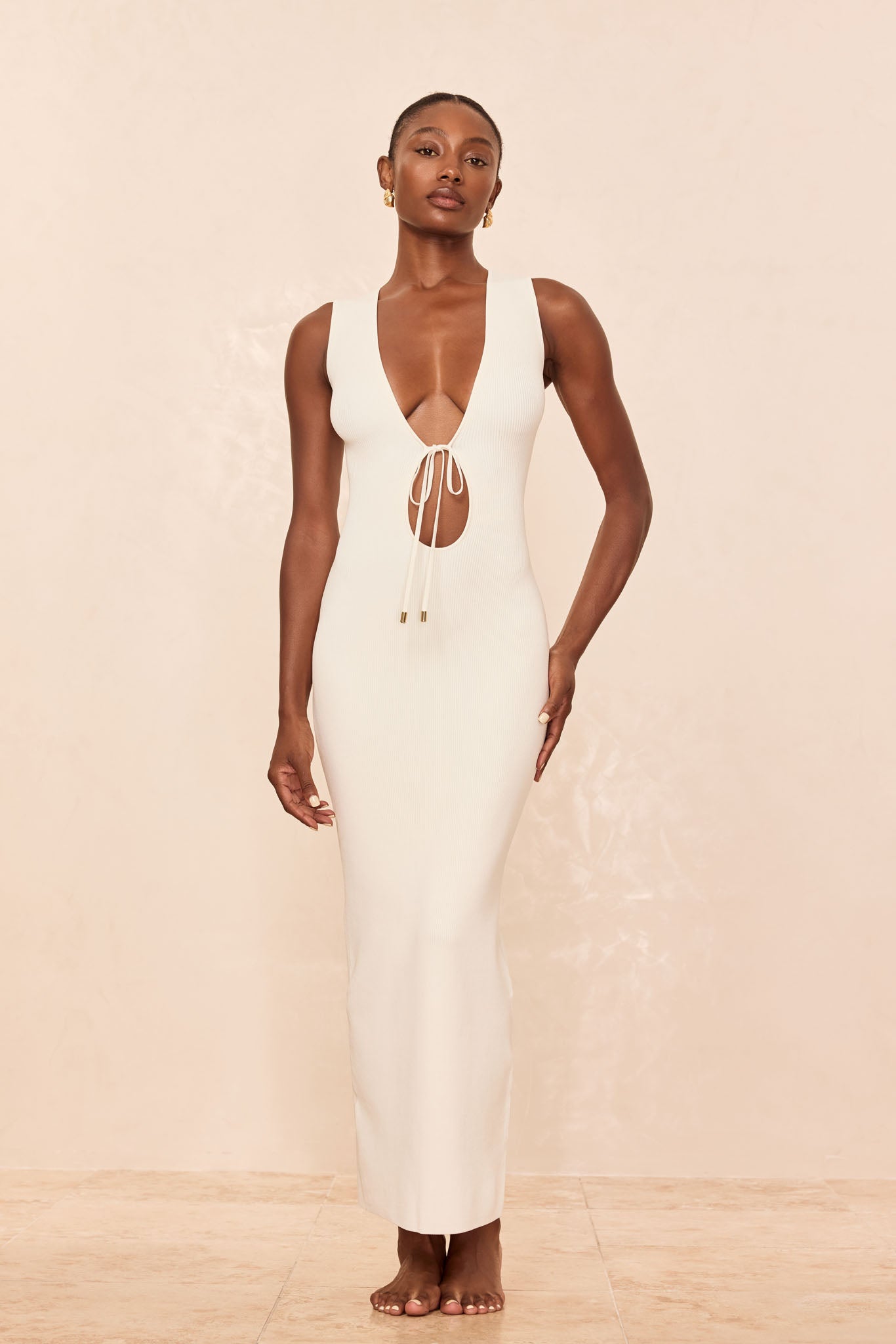 Sleeveless cream ribbed maxi dress with deep V and tied keyhole front, shown on a standing model against a neutral background.