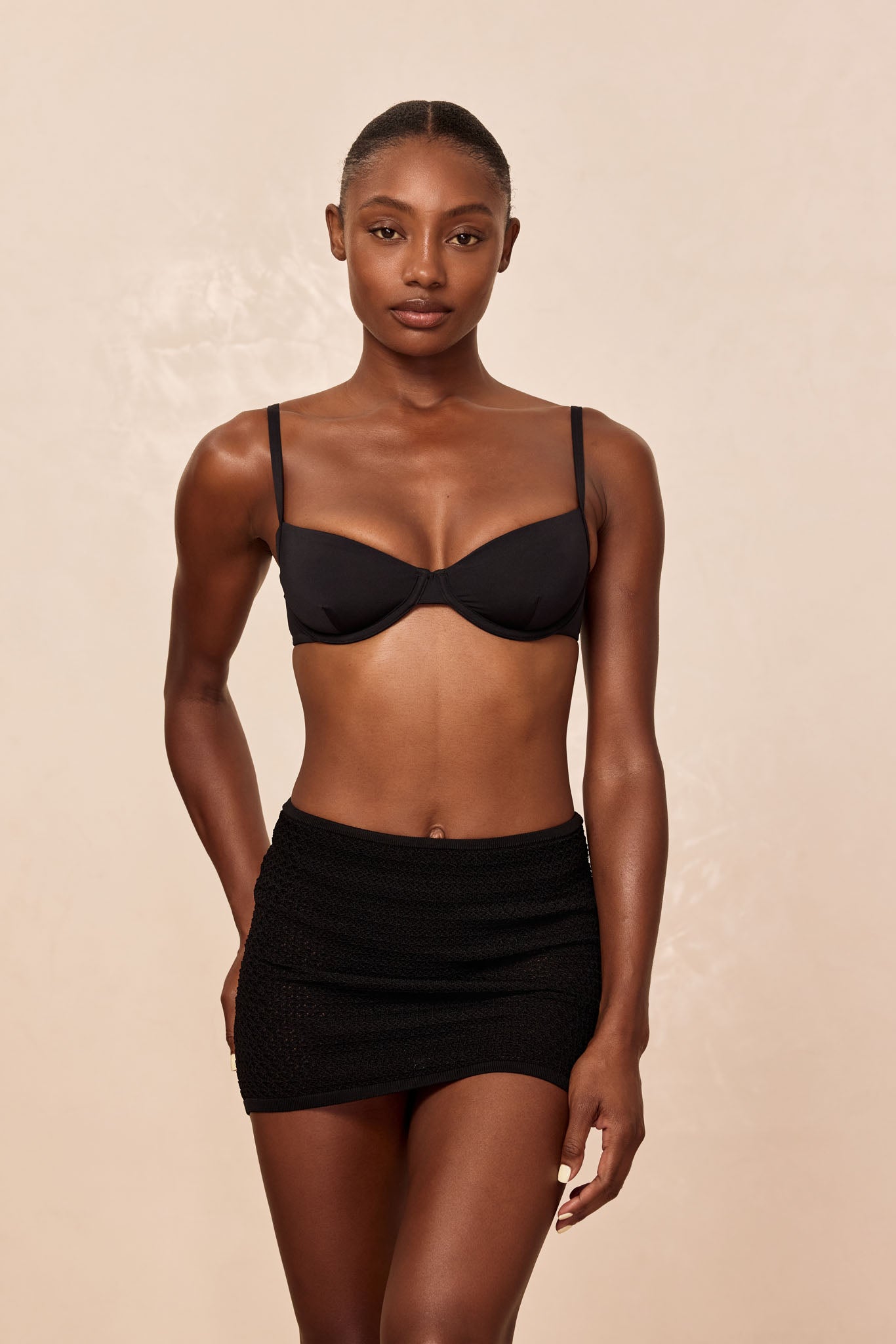 Model wearing a black underwire bra and black textured mini skirt, frontal pose against a beige background.