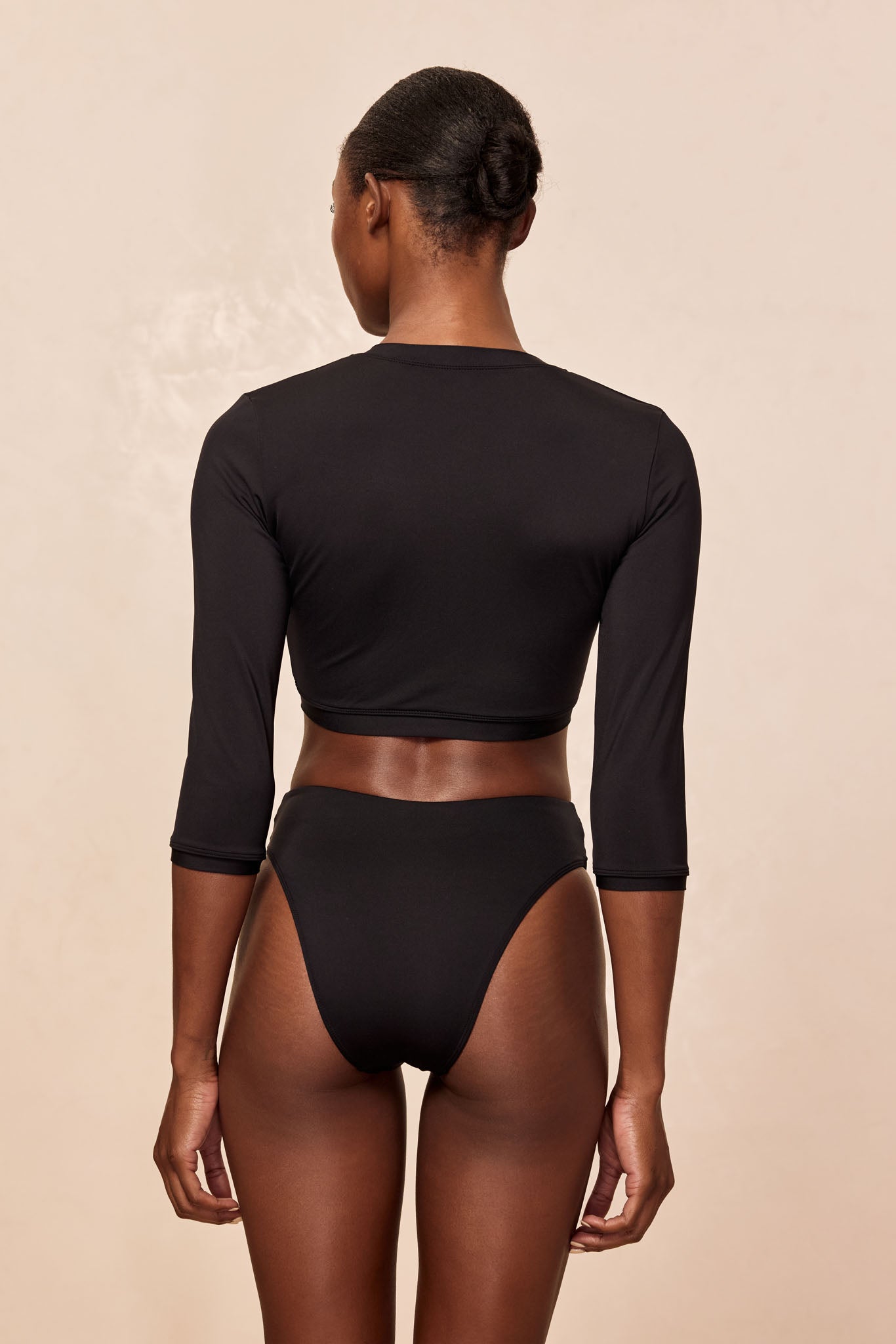 Rear view of model wearing black long-sleeve crop swim top and high-cut bikini bottoms on neutral background.