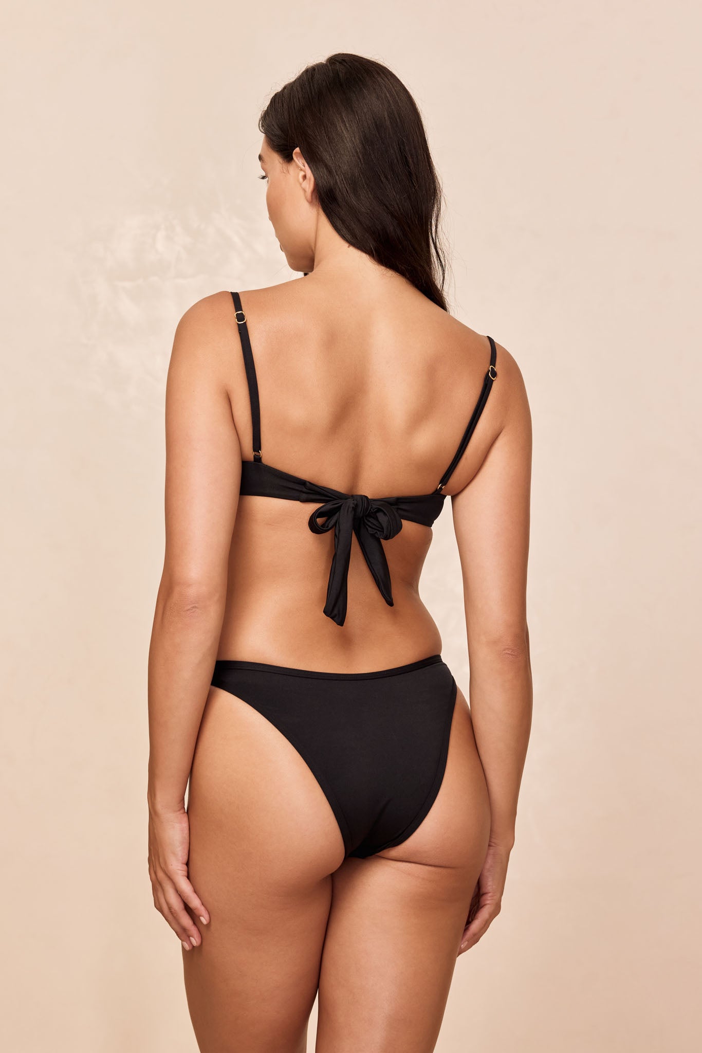 Back view of woman wearing black tie-back bikini top and high-cut bikini bottoms on neutral background.