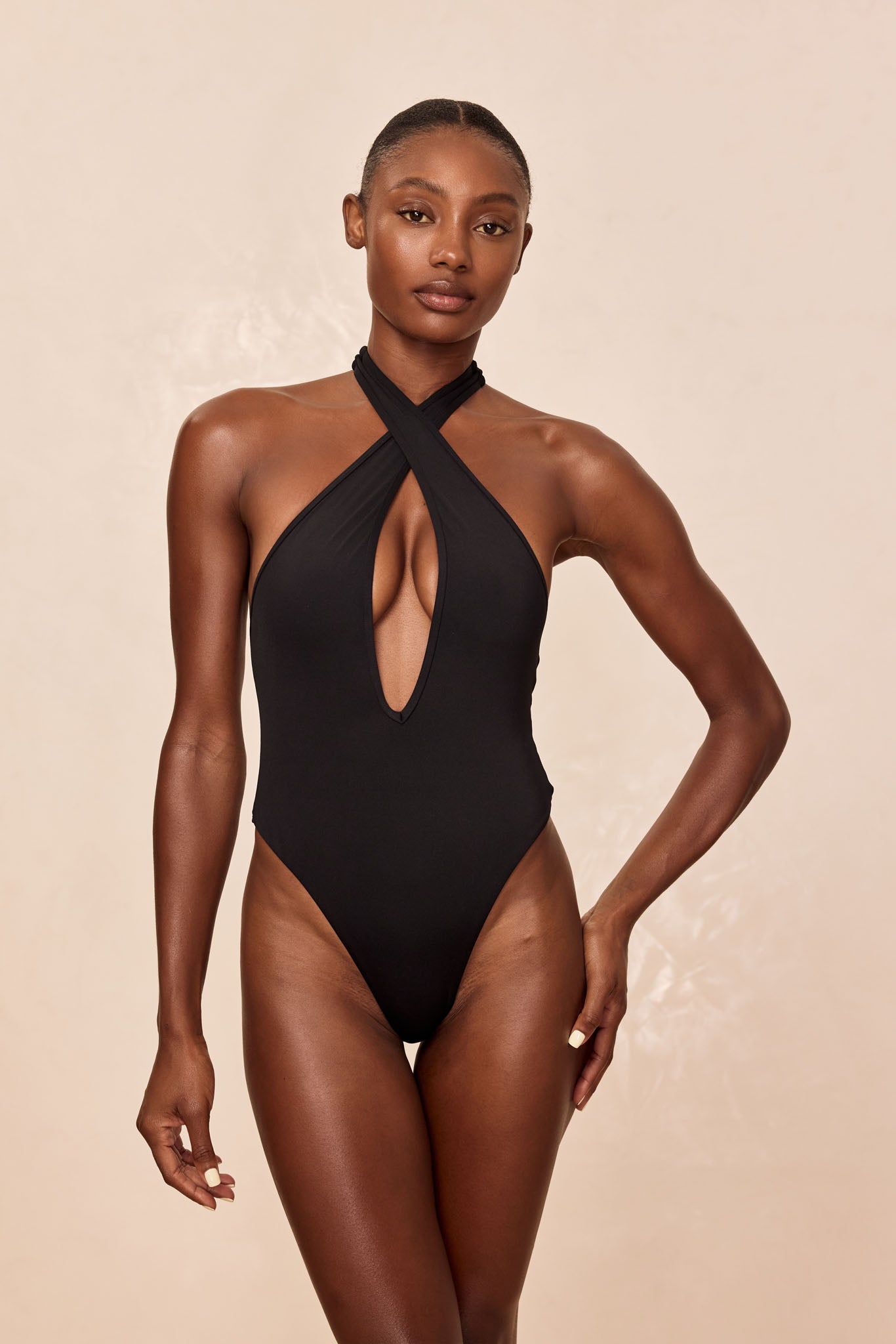 Model wearing black one-piece swimsuit with crossed halter straps and deep keyhole front, standing against beige background.