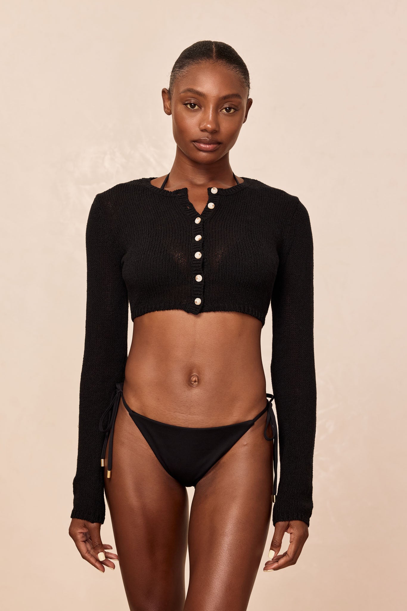 Model standing front-facing wearing a black cropped buttoned sweater and black tie-side bikini bottoms.