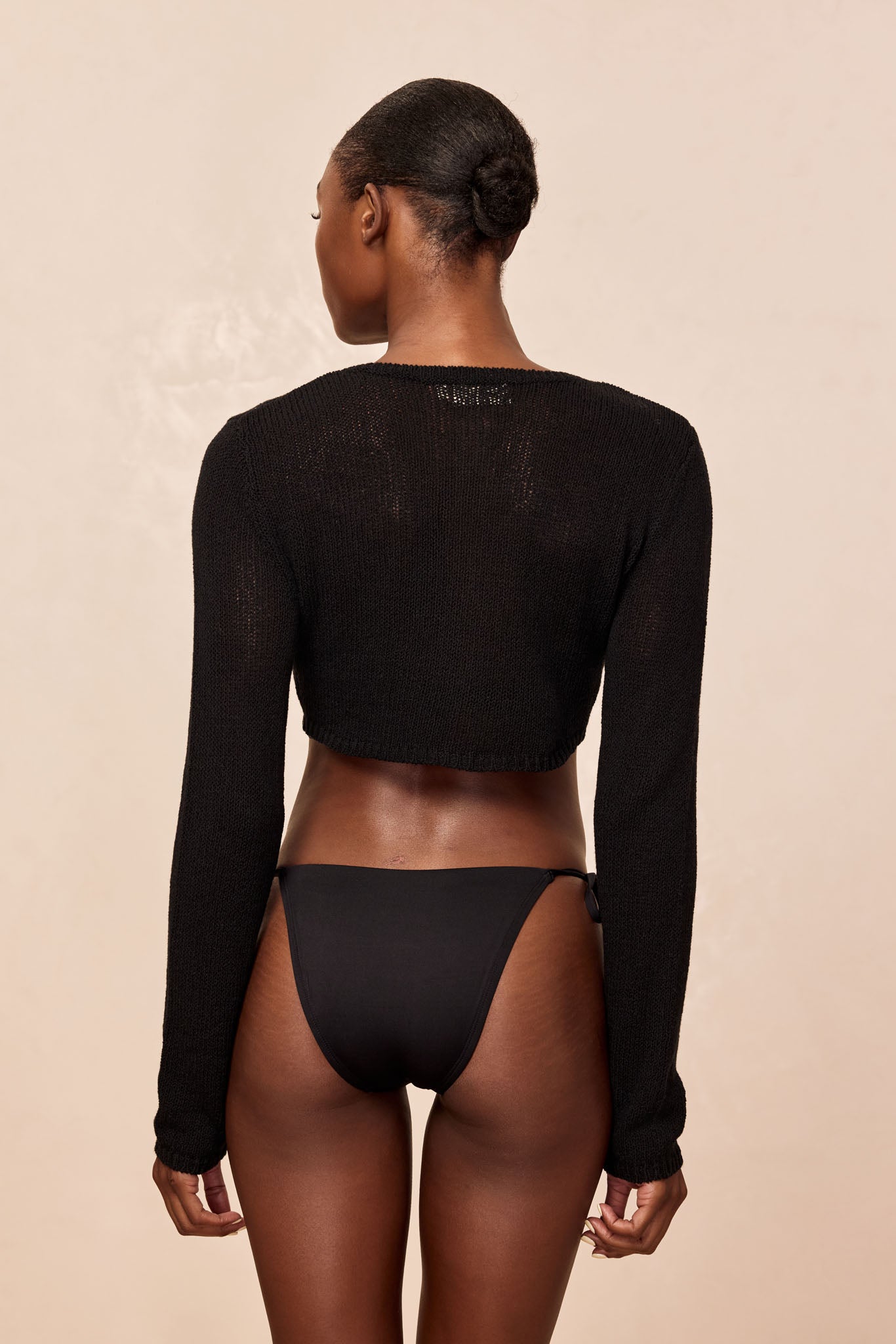 Back view of woman in a low bun wearing a black cropped knit sweater and black bikini bottoms on beige background.