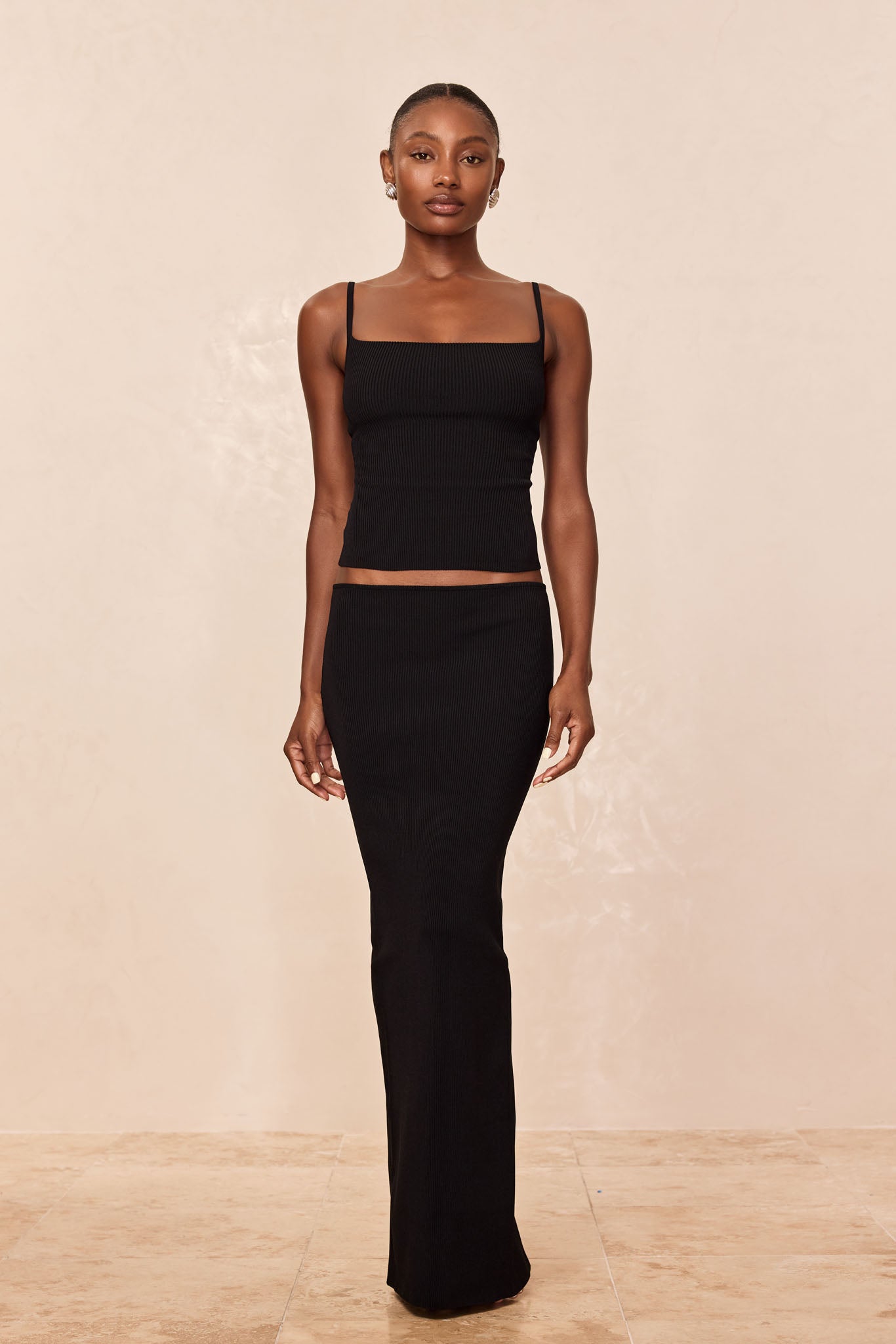 Woman wearing black ribbed square-neck cropped tank top and high-waisted long black skirt, standing against beige background.